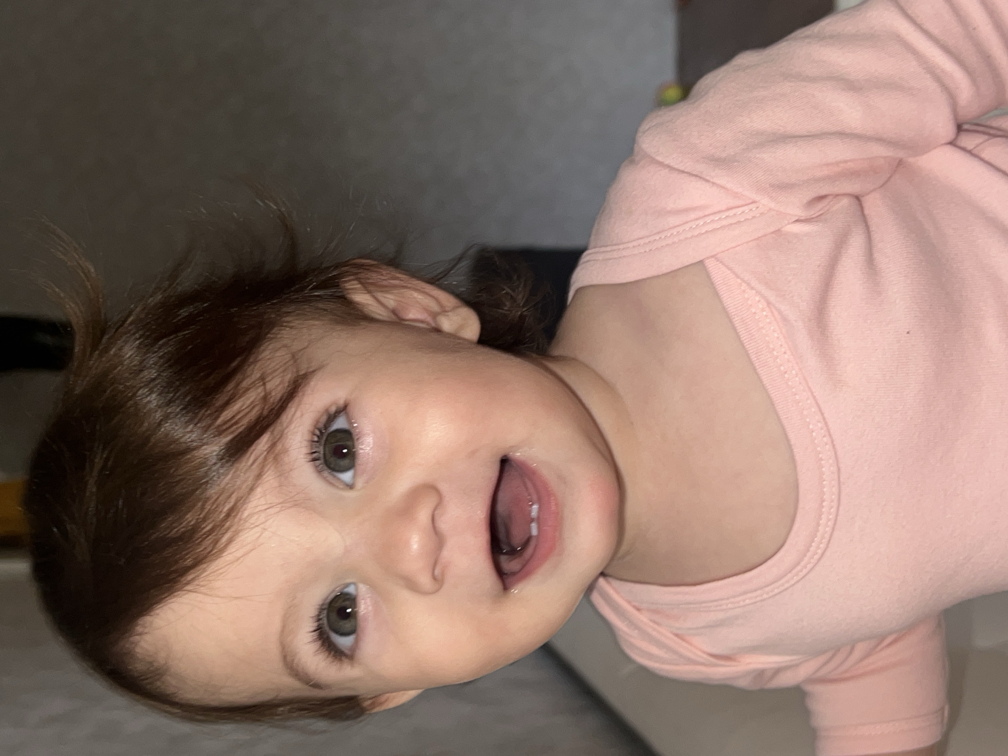 A close-up, slightly rotated, indoor portrait captures a young child with fair skin, dark brown hair, and light-colored eyes. The child is wearing a light pink, long-sleeved garment. The child's mouth is open, revealing teeth. The background is blurred, with a gray wall visible. There is nothing to suggest that the photo was taken in Elena, Bulgaria.