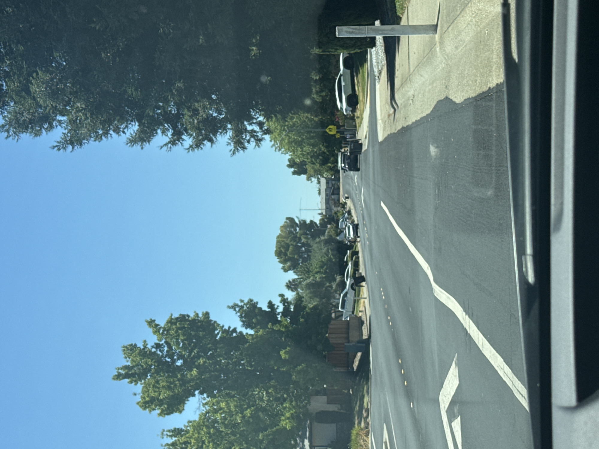 This image depicts a street scene in Roseville, United States of America, on a clear, sunny day. The perspective is from inside a vehicle, looking forward. The road is asphalt, with clear white lane markings, including directional arrows for turning left. Several cars are parked or driving on the right side of the road. On the left side of the road, there are houses and mature trees. On the right side, there is a sidewalk, more trees, and a yellow school crossing sign. The sky is a vibrant, cloudless blue. The overall impression is of a quiet, suburban residential street on a pleasant day.