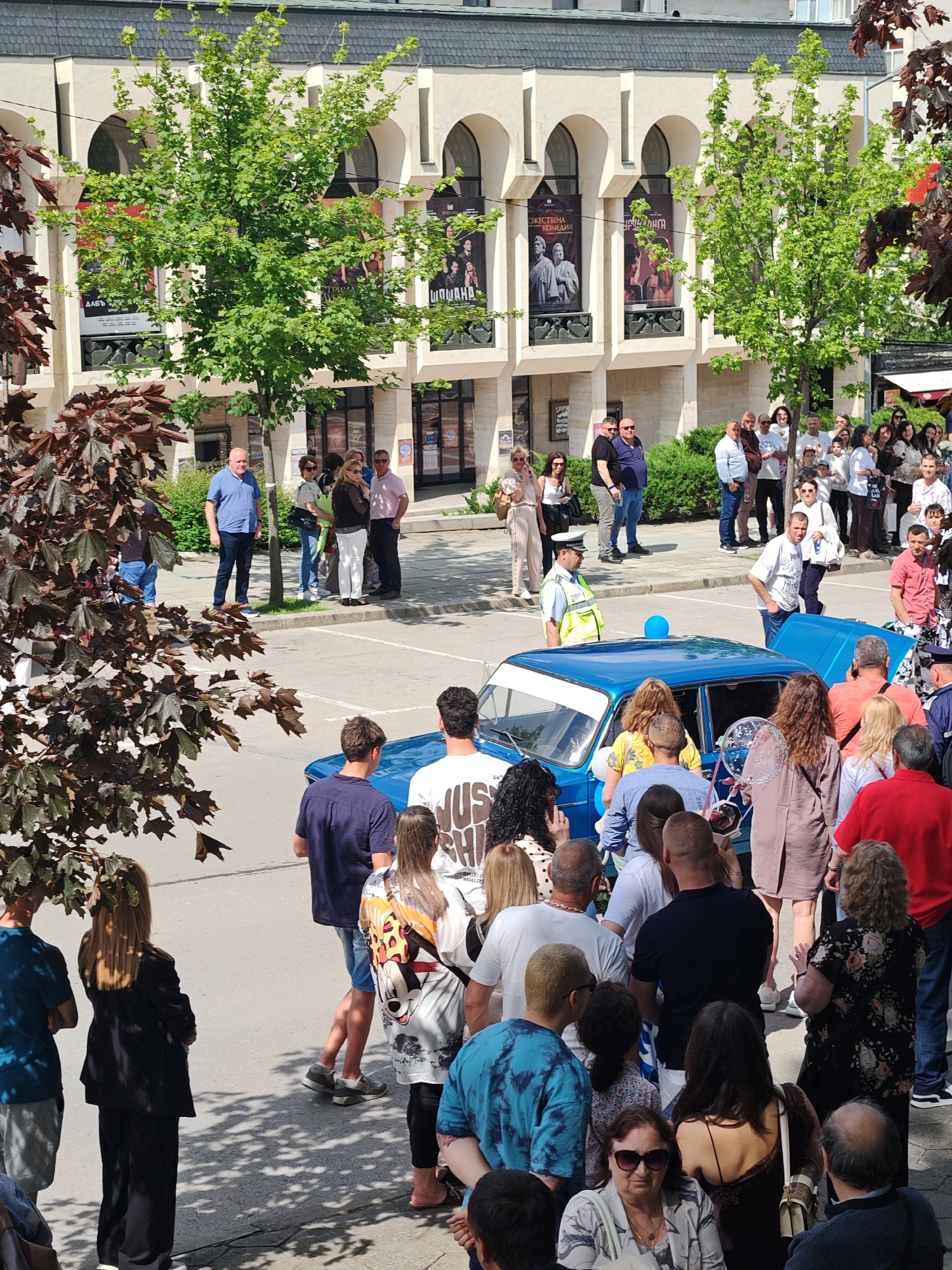 The image depicts a crowd of people gathered around a blue car in the city of Shumen, Bulgaria. The location appears to be in front of the Vasil Drumev Drama and Theatre. The crowd consists of people of various ages and attire. A police officer is standing near the car, and the car has its hood open. Some individuals appear to be interacting with the car, while others are observing. The setting is outdoors, with trees and buildings visible in the background. Banners related to performances are present on the theatre building.