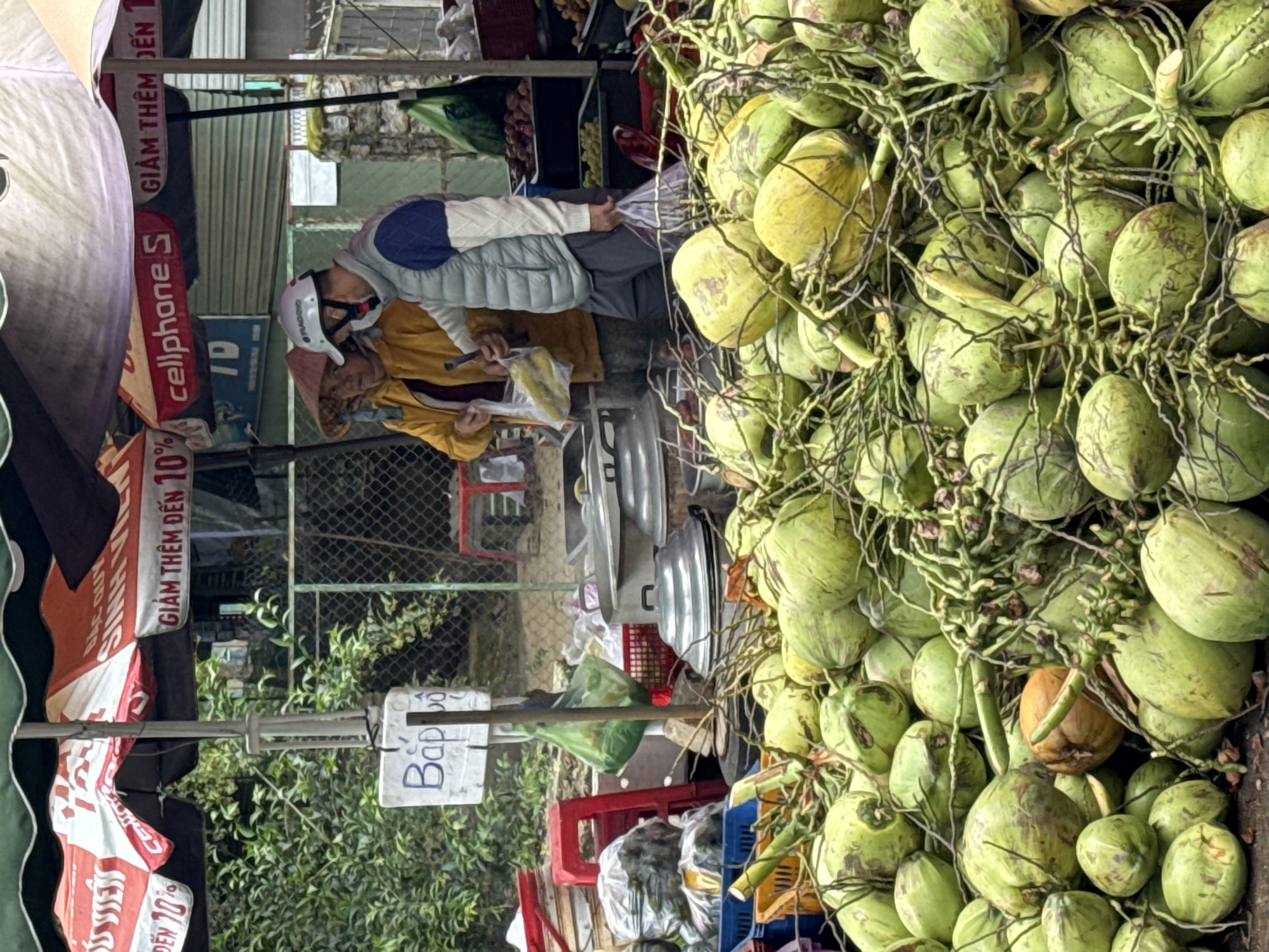 This media file captures a bustling street market scene, likely in Lâm Viên - Đà Lạt, Vietnam. The right side of the image is dominated by a large, fresh pile of green coconuts, some still attached by their stems, suggesting a fruit stall. Among the green coconuts, one brownish-yellow one is visible.
In the center-left, two individuals are interacting. One person, possibly a customer, wears a light-colored vest, a blue and white long-sleeved top, and a white helmet with "PIAGGIO" written on it. Facing them is a vendor, wearing a yellow long-sleeved shirt and a traditional Vietnamese conical hat (nón lá). The vendor appears to be holding a tool, possibly a coconut husker or machete, and a plastic bag or another coconut. Behind the vendor, several large metal pots or containers and plastic crates are visible, indicating other goods or food items being sold.
The setting is outdoors, with natural light and glimpses of foliage and a wire fence in the background. Buildings with metal roller shutter doors are also faintly visible.
Several banners and signs add to the market atmosphere. Visible text includes "GIẢM THÊM ĐẾN 10%" (Get an additional 10% discount), "cellphoneS", "ĐẾN 70%" (up to 70%), "10%", "SINH QUÝ", and "BAC QUY" on various promotional materials. A smaller sign hanging on a pole clearly reads "Bắp Bo". The activity depicted is a typical market transaction between a vendor and a customer.