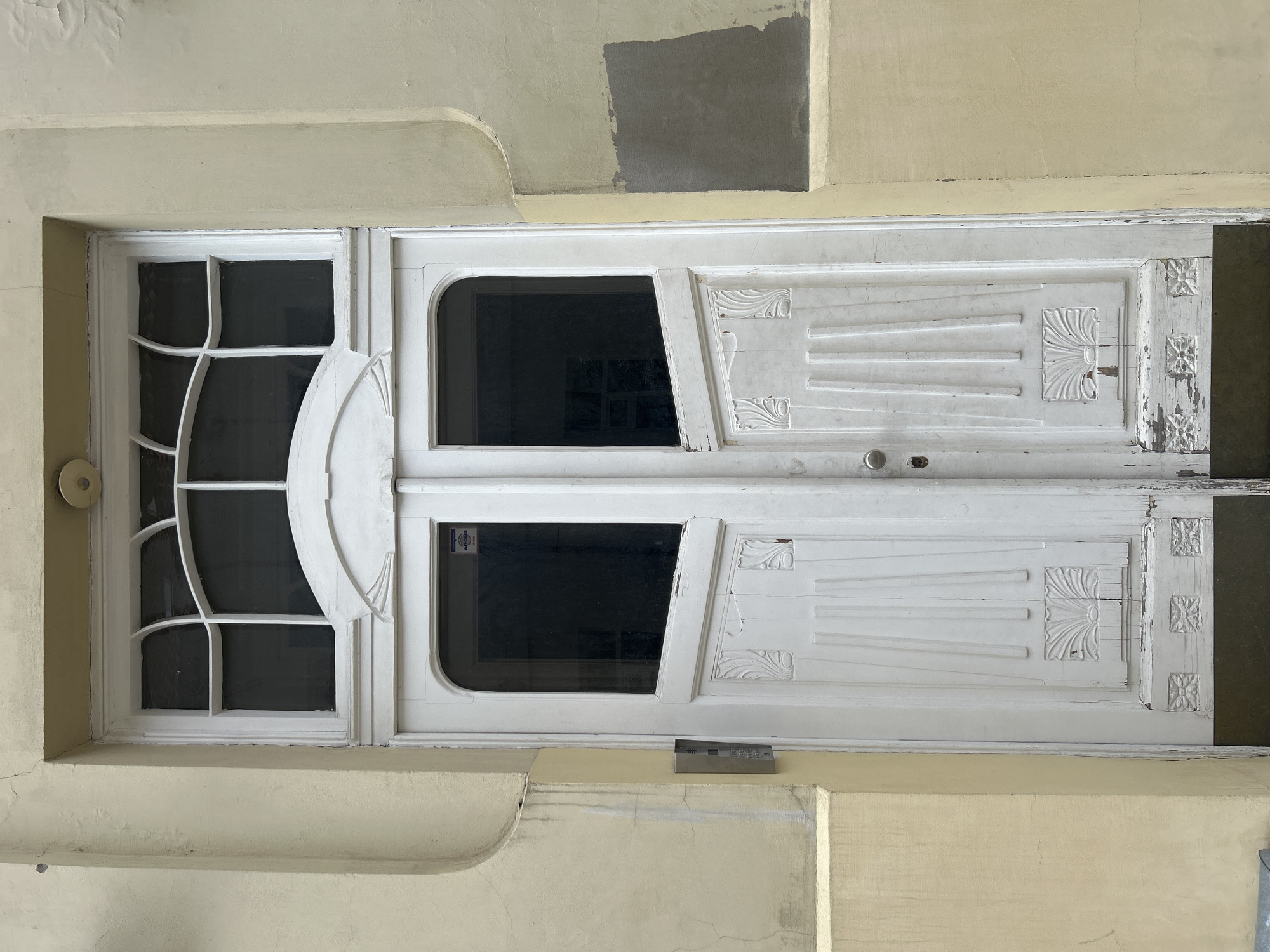 The image shows the front of a weathered white double door set into a pale yellow wall. The door has glass panels at the top and decorative paneling on the lower half. Above the door is a large window with curved mullions. To the left of the door, mounted on the wall, is a silver intercom unit. The wall shows signs of age and wear, with some discolored patches and cracks. The scene is likely an entrance to a residential building or a commercial establishment. The location is identified as Vilnius, Lithuania.