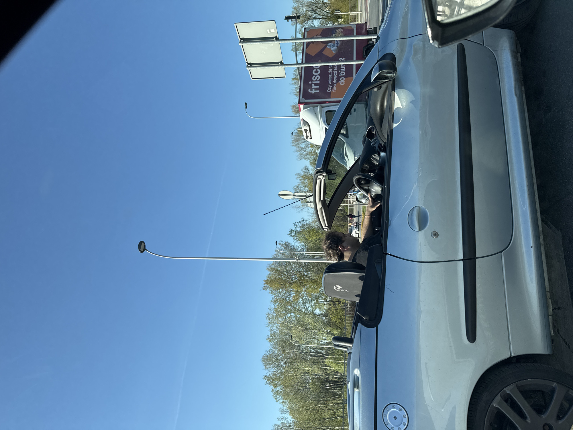 A silver convertible car with the top down is positioned on a paved road. The driver is visible, wearing a black shirt and glasses. In the background are trees, streetlights, and a large advertisement for "frisco.pl". The text on the advertisement is in Polish. The scene is set during the daytime under a clear, blue sky. Based on the frisco.pl advertisement, the location is Warsaw, Poland.