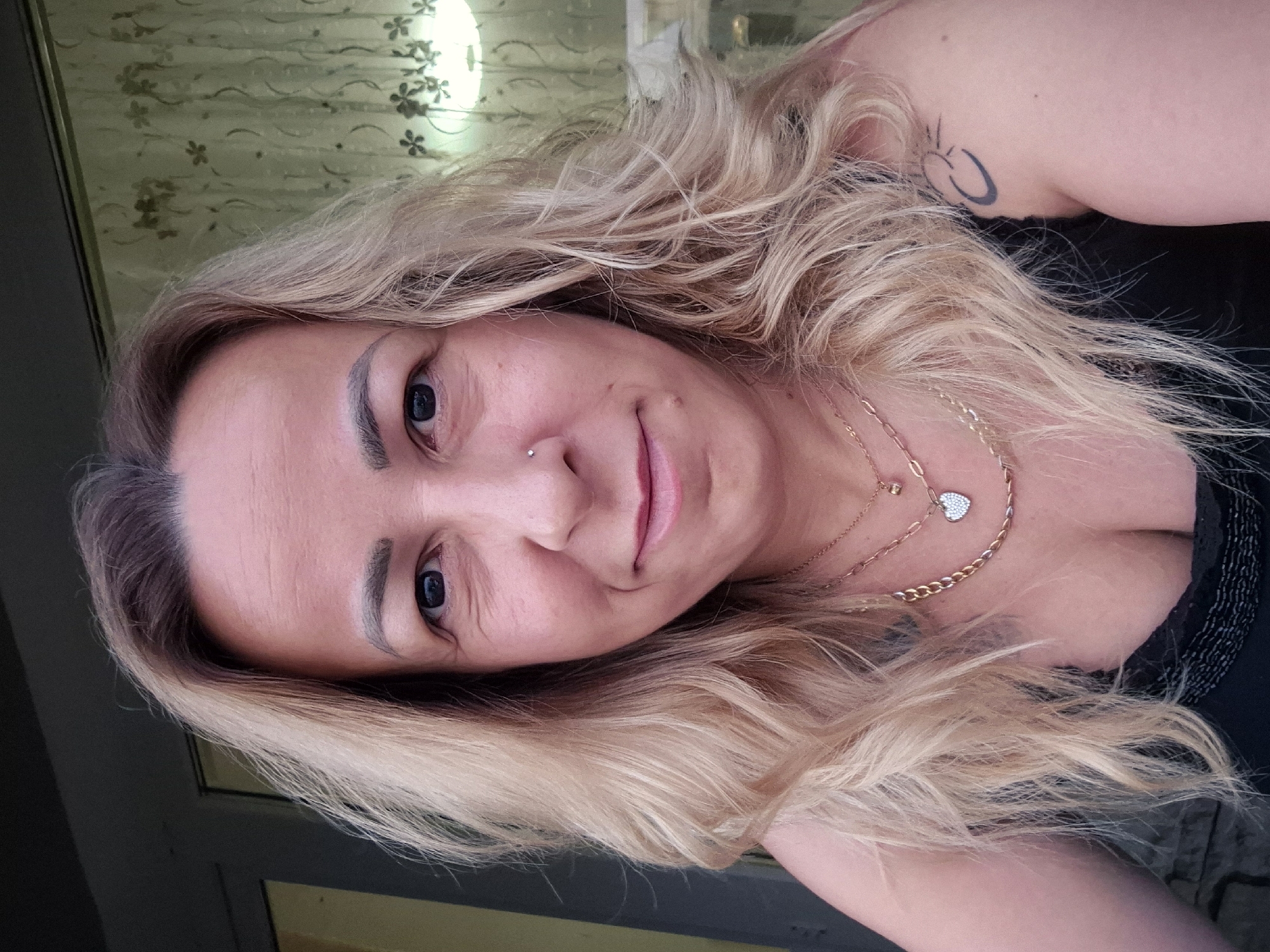 A medium shot captures a woman from the chest up, facing the camera directly. She has blonde, wavy hair that falls past her shoulders. Her eyes are dark, and she has a small nose stud. She is wearing two necklaces, one a gold chain with a small heart pendant covered in what appears to be rhinestones, and another gold chain with a simpler heart charm. Her right shoulder is visible, showing a tattoo of a crescent moon with a sun in the center. She is wearing a black top with some embellishment around the neckline. The background is out of focus, with a blurred curtain that has a floral pattern and a light fixture visible. The overall lighting suggests an outdoor setting, possibly during the day.