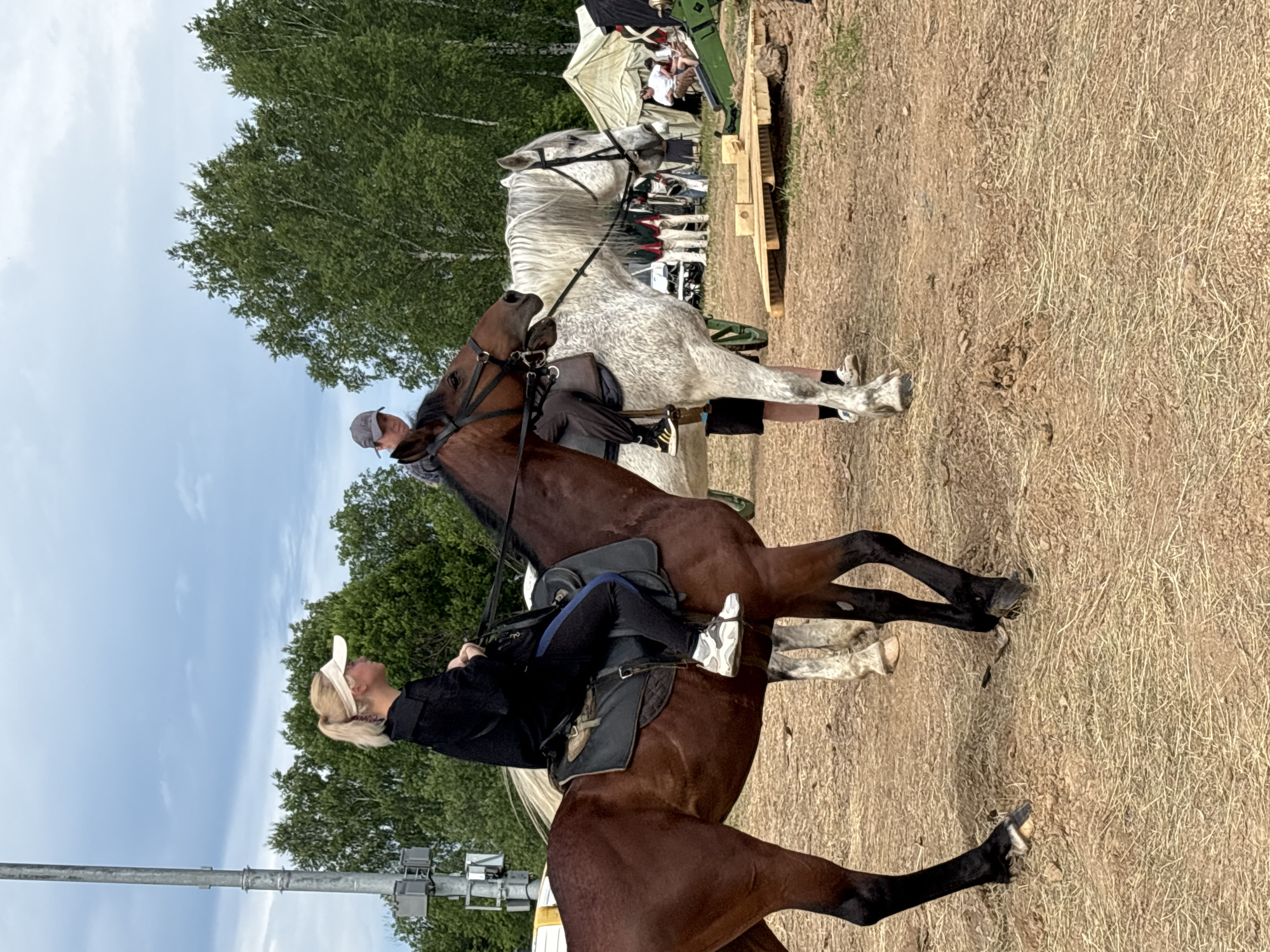 In the city of Bornosovo, Russia, a horse riding event is underway. Two people are mounted on horseback. The person on the left, a woman with blonde hair tied in a ponytail, wears a black top, dark pants with a blue stripe, and white sneakers. She is on a brown horse, its head turned to the right. To the right, a person wearing a grey baseball cap and a grey checkered shirt rides a white speckled horse. The horse's head is also turned to the right, with its mouth slightly open. In the background, several people are gathered near tents. A green cannon-like object sits on a wooden platform in the foreground. The ground is covered in dry straw and dirt. Trees form a backdrop against a cloudy sky.