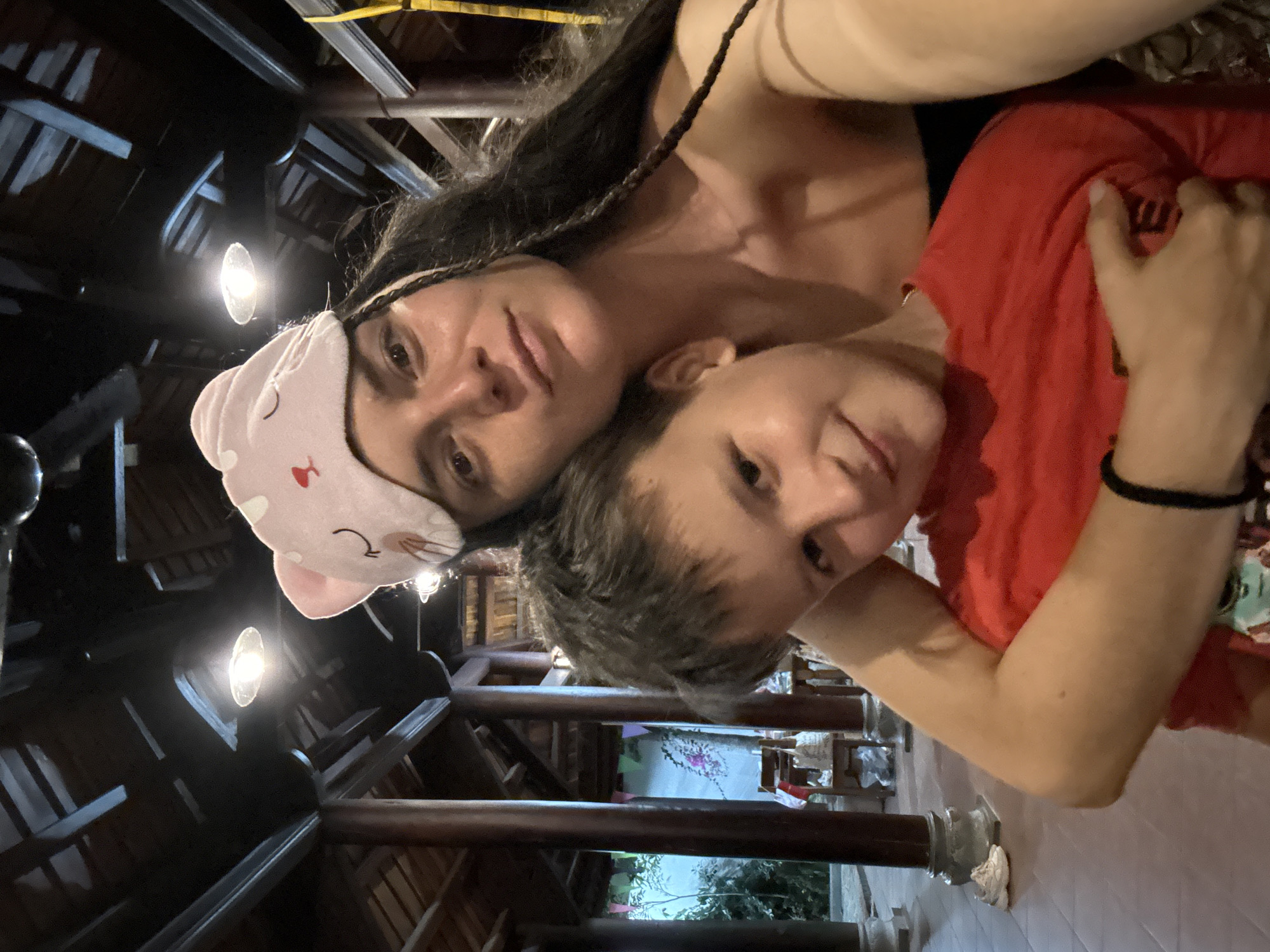 A selfie shows a woman and a boy, embraced closely, in an indoor, presumably commercial, space in Mũi Né, Vietnam. The woman, with long, dark, partially braided hair, wears a black tank top and a pink cat-ear sleep mask pushed back on her forehead. The boy, with short, brown hair, wears a red t-shirt. He has his arms around her. The background features interior architecture with dark wood beams and supports, and hanging lights. In the far background, a garden and furnishings are faintly visible through the posts. The photo is oriented with the subjects in the upper left and right, and the architectural details in the lower half.