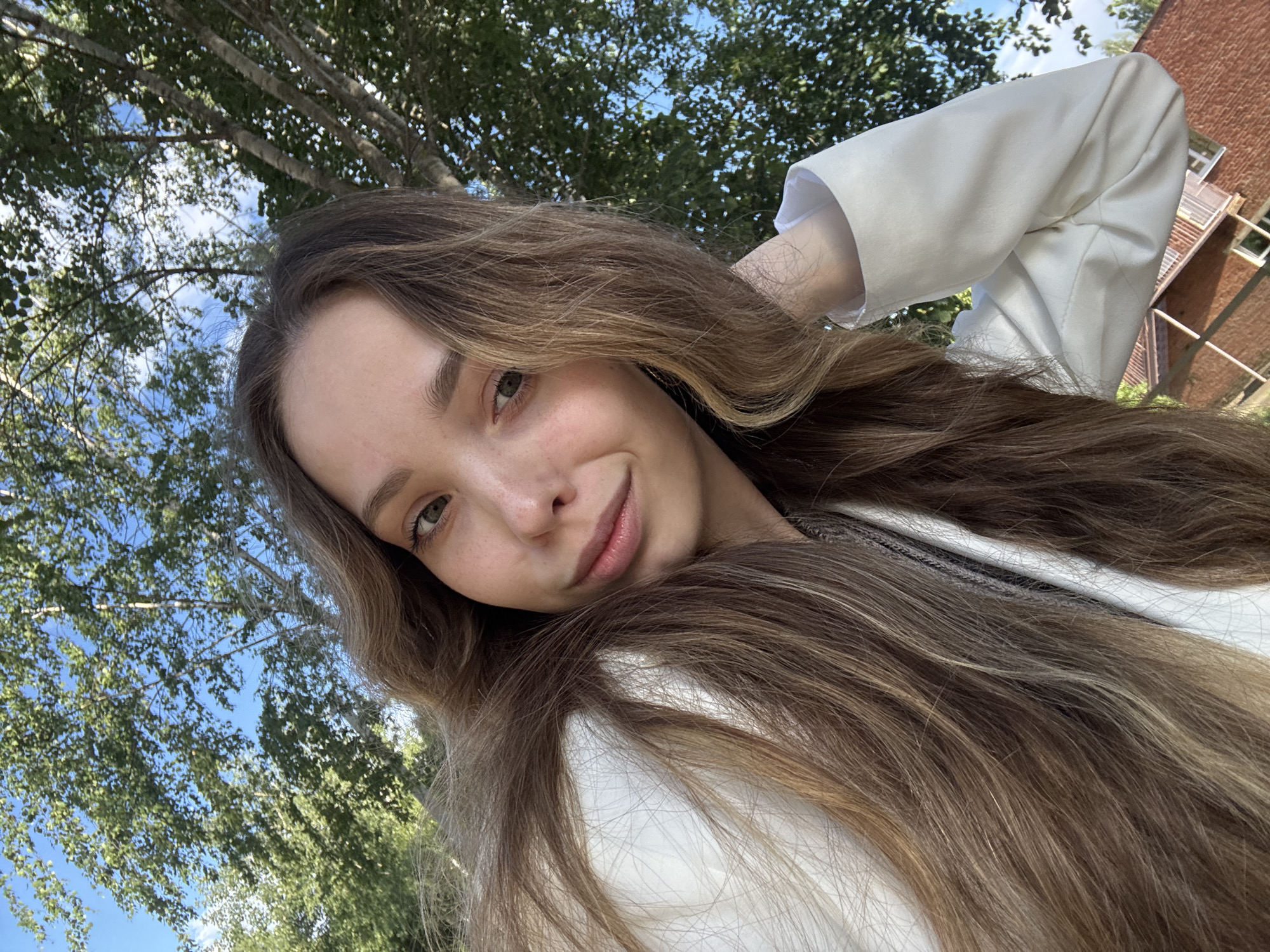A young woman with long, wavy brown hair is captured in a selfie taken outdoors. She is wearing a white blazer. Her head is tilted, and her right arm is bent, with her hand resting behind her head. Her gaze is directed towards the camera, and she has a slight smile. The background consists of green trees and a clear blue sky, suggesting a sunny day. To the right of the frame, a brick building is partially visible, indicating an urban or semi-urban setting. The lighting suggests it is daytime, likely mid-morning to afternoon.