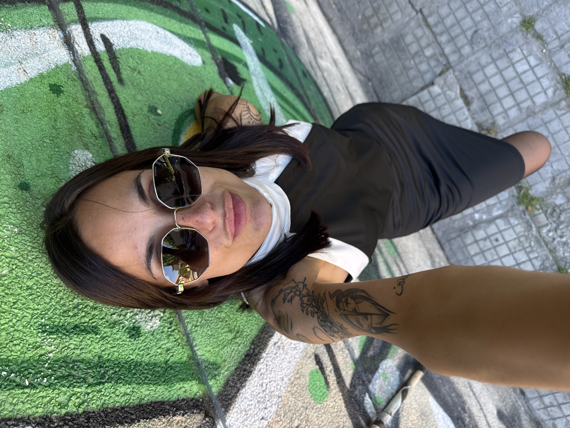 A woman lies on a sidewalk next to a graffiti-covered wall in what appears to be the city of Pleven, Bulgaria. She is wearing sunglasses and a black dress with a white collared shirt underneath. She has brown hair and visible tattoos on her arms. The sidewalk is tiled and the graffiti is primarily green and white. The woman is positioned in a way that suggests she may be taking a selfie or having someone take her picture from an overhead angle.