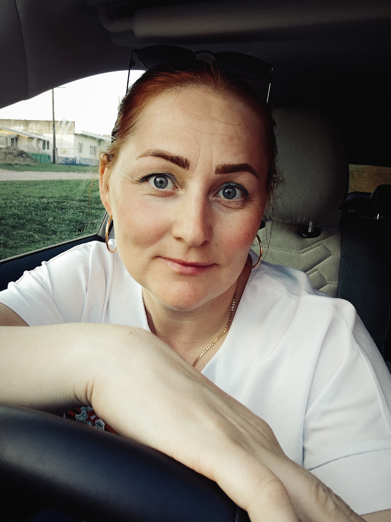 The image is a close-up, eye-level selfie of a woman inside a car. The woman has fair skin, reddish hair pulled back, and bright blue eyes. She is wearing a white top, gold hoop earrings, and a gold necklace. Sunglasses rest on top of her head. Her left arm is resting on the steering wheel.
Through the car window, one can see a rural or suburban landscape. There is green grass and some buildings, including a white building with a blue fence. This is in the city of Фоки, Russia. The lighting suggests it is daytime.