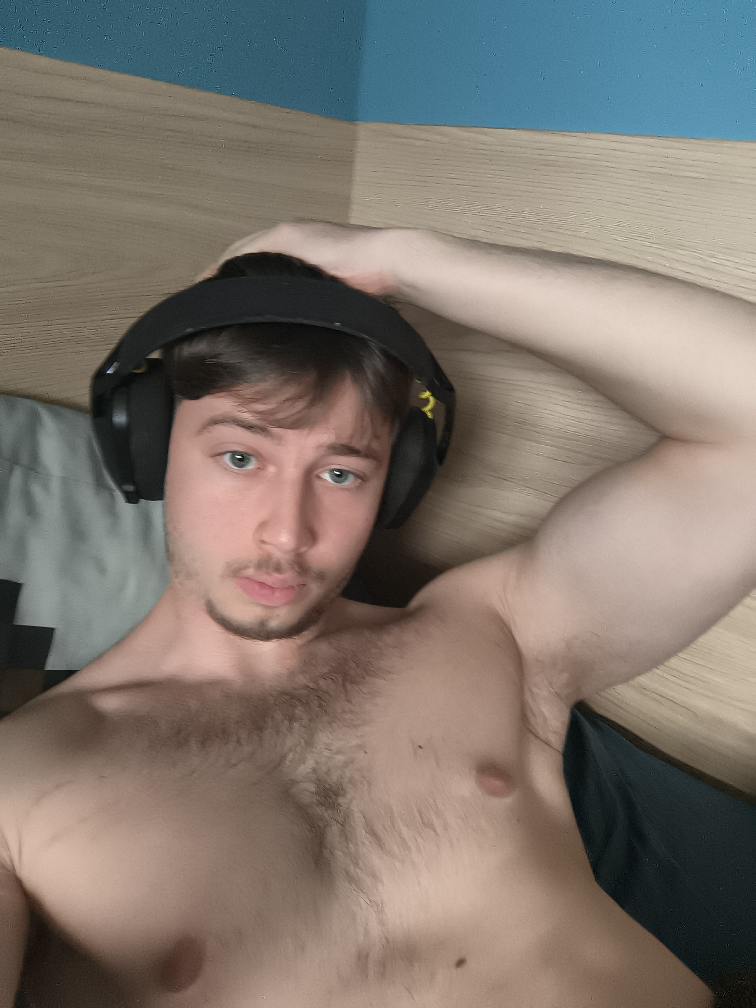 A shirtless man with blue eyes and brown hair is wearing black headphones. He is lying down on what appears to be a bed with a grey pillow and a dark teal sheet. His right arm is raised, with his hand on his head. Behind him is a wooden headboard and a light blue wall. There is no indication of the location being Sofia, Bulgaria.