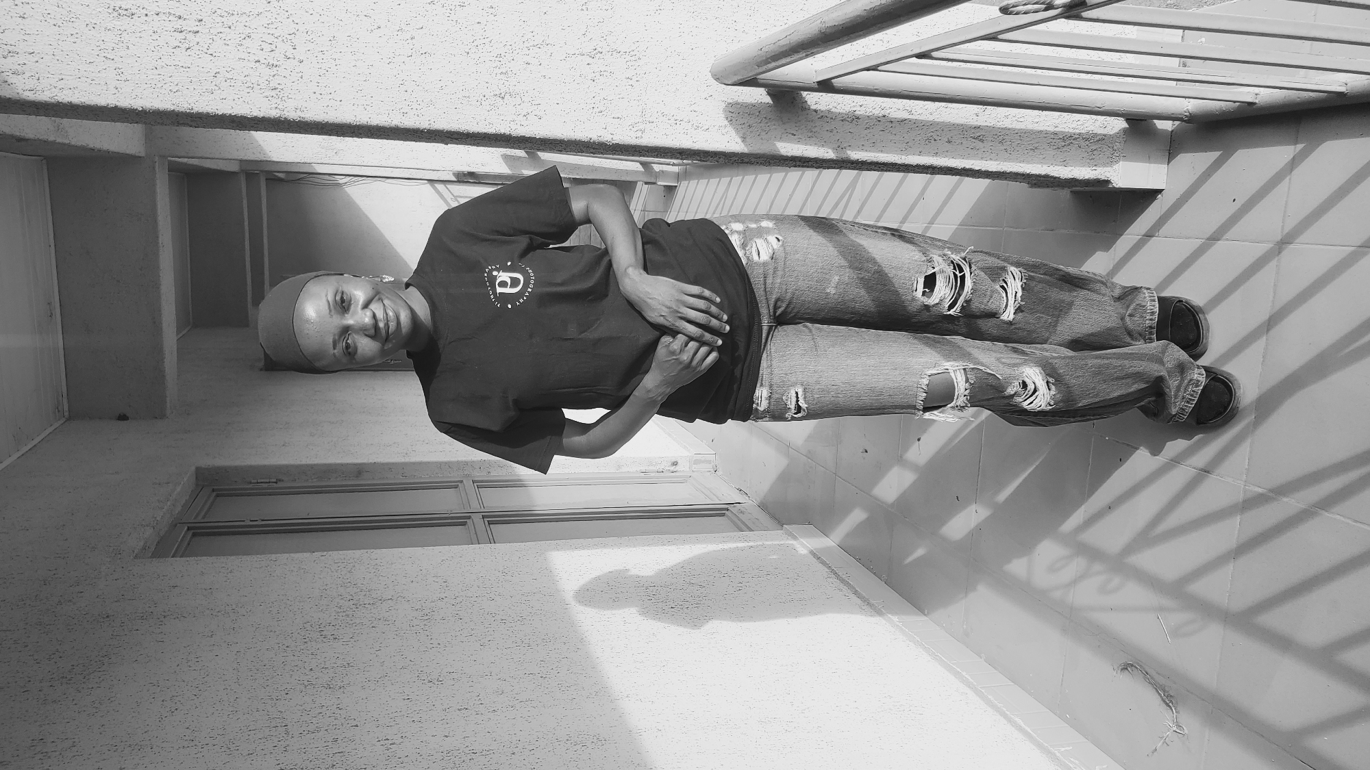 A person stands in a black t-shirt and ripped jeans on a tiled surface, likely a balcony, in Bukuru, Nigeria. The person has a head covering and their hands are clasped at their waist. Behind them is a window and above them is a metal railing. The scene is in black and white.