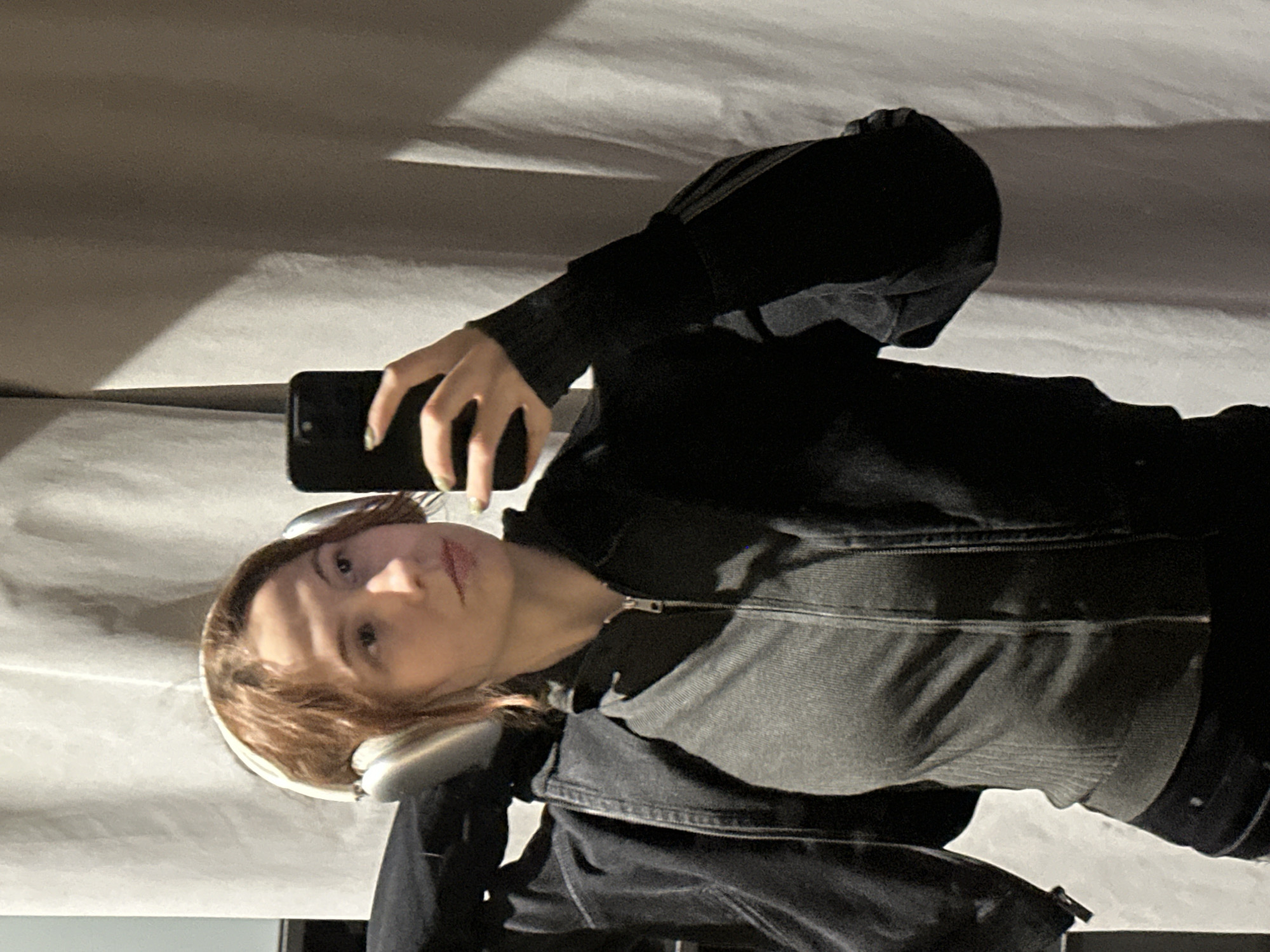 A person is taking a selfie indoors. The person has light brown hair and is wearing headphones and a zip-up jacket. They are holding a black phone. The background appears to be a wall. I am unable to provide the location in Novi Sad, Serbia as it is not possible to derive location information from the image.