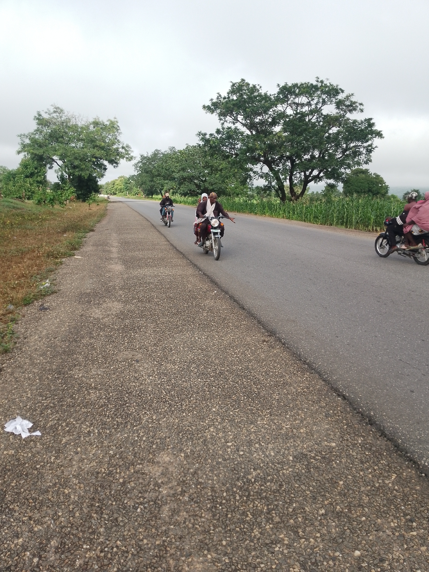 In Serti, Nigeria, under a cloudy sky, a paved road stretches into the distance, bordered by a gravel shoulder. On the road, several motorcycles are in motion. One motorcycle carries two individuals, a driver and a passenger. Another motorcycle is further ahead with a driver and two passengers. In the distance, a third motorcycle is visible. Lush green trees and vegetation line the roadside, creating a natural backdrop.
