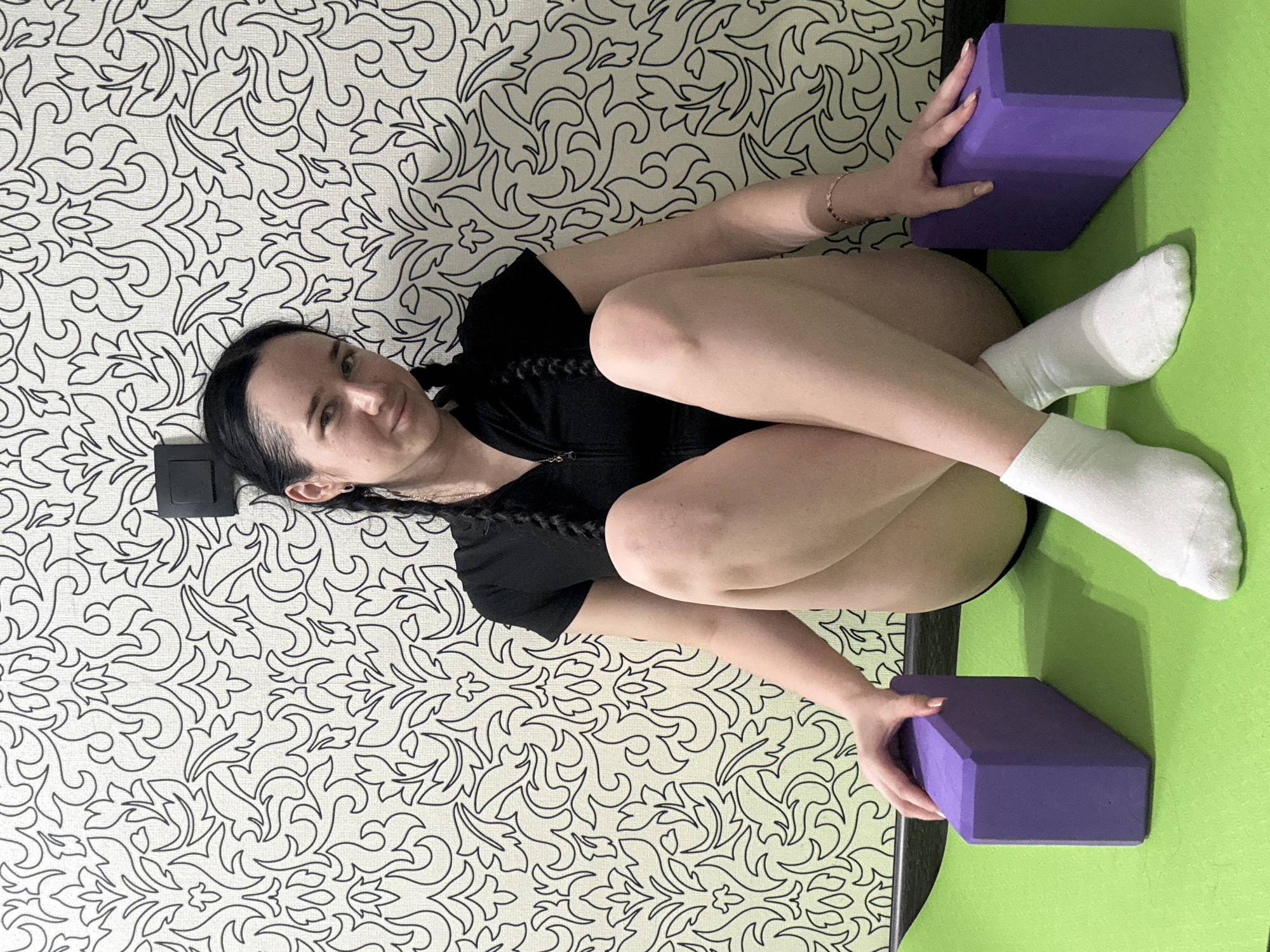 A young woman with dark hair in braids sits on a bright green yoga mat in front of a white and black patterned wallpaper. She is wearing a black athletic top and white socks. Her legs are crossed, and she is holding two purple yoga blocks, one on either side of her. The room appears to be indoors, likely a home or a studio, with a light switch visible on the wallpaper. There is no indication of the location being Novosibirsk, Russia.