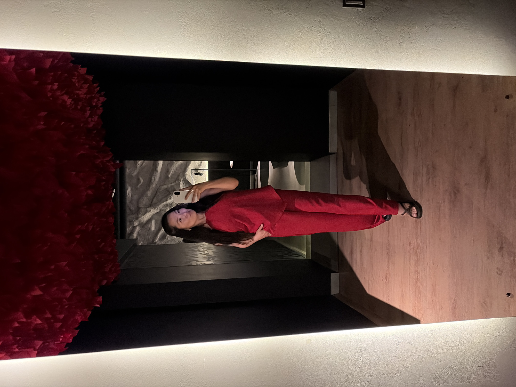 The image is a selfie taken by a woman in a room with unique architectural elements. The woman is wearing a red pantsuit and black sandals, holding a smartphone to take the picture. She stands in front of a wall with a large, red floral art piece on the left and a black divider. Behind her, there's a bathroom with marble-patterned walls. The floor appears to be wooden. The room is illuminated by strip lights along the top of the walls. There is no specific information from the picture about the city of Chaikovsky, Russia. The image shows a residential setting, possibly a hotel room.