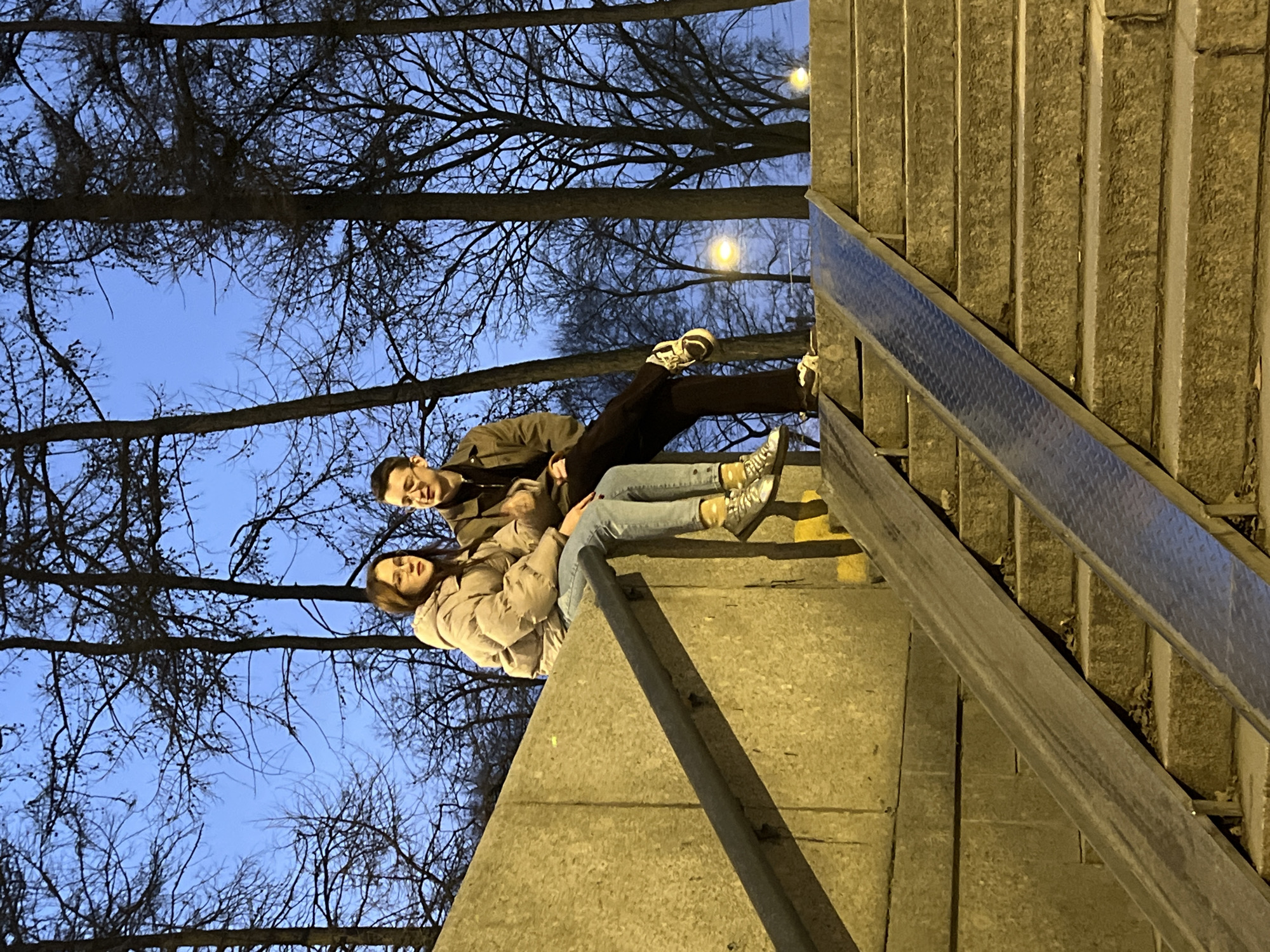 Two individuals are seated on a concrete wall that forms part of a staircase structure in an urban park within Saint Petersburg, Russia, under dusk or night conditions. The background shows numerous tall, bare trees silhouetted against a deep blue sky, with two distant streetlights emitting a warm glow.
The female, positioned on the left, wears a light-colored (beige or tan) puffy winter jacket, light blue jeans, and reflective silver sneakers over yellow socks. She has dark hair, wears glasses, and appears to be speaking, with her mouth slightly open and her gaze directed to her right. The male, seated next to her on the right, wears a dark jacket, dark trousers, and light-colored sneakers. He also wears glasses and is looking towards the female, exhibiting a slight smile.
Below the concrete wall, a wide concrete staircase descends towards the right. Two metal ramps are integrated into the staircase: the left ramp is smooth, and the right ramp features a diamond plate pattern. A dark metal handrail runs diagonally on the left, parallel to the concrete wall. Yellow markings are visible on the concrete structure behind the handrail. The scene is illuminated by artificial light, imparting a yellowish hue to the concrete surfaces.