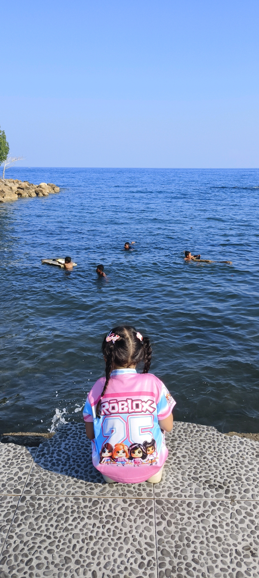A young girl with pigtails, wearing a pink and blue Roblox shirt with cartoon characters and the number "25", is kneeling on a textured grey stone pathway. She is facing away from the camera, looking out at a body of water that appears to be the Raja Ampat Islands, near Waisai, Indonesia. The water is calm and blue, with several children swimming in the distance. One child is on a flotation device. The shore to the left is lined with rocks and a small tree. The sky is clear and bright blue. The lighting suggests it is daytime, likely mid-morning or early afternoon.