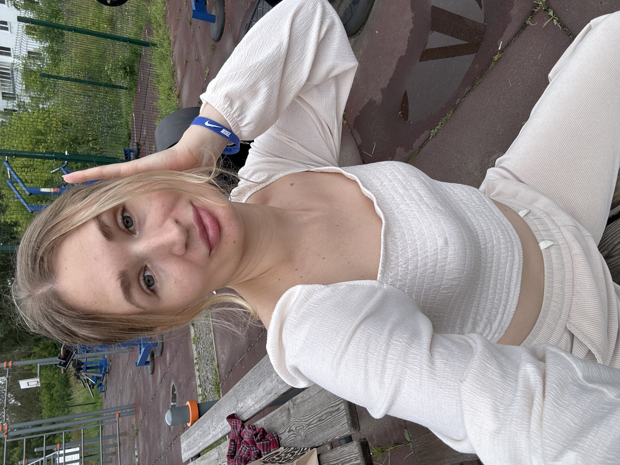 A young, blonde woman is sitting on a wooden bench at an outdoor fitness equipment area. She is wearing a white, long-sleeved, off-the-shoulder top and light-colored pants. On her left wrist, she wears a blue Nike bracelet. The background includes various pieces of outdoor exercise equipment, a chain-link fence, and some trees and buildings. The ground is paved with red rubber tiles, some of which are wet. The location is an outdoor fitness park in Chaikovsky, Russia.