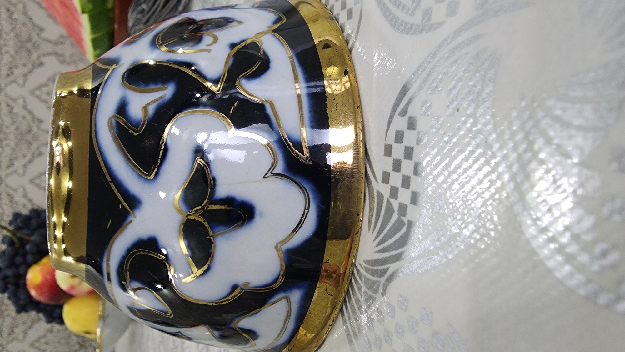 The image shows a close-up of a ceramic bowl with a white base, decorated with dark blue and gold floral patterns. The bowl has a thick gold rim. It is placed on a white tablecloth with a subtle checkered and fan-like pattern. In the blurred background, on the left, are some fruits including grapes, apples, and a lemon. On the right, a slice of watermelon is visible. The scene appears to be set indoors, possibly on a table in a home or restaurant in Margilan City, Uzbekistan.