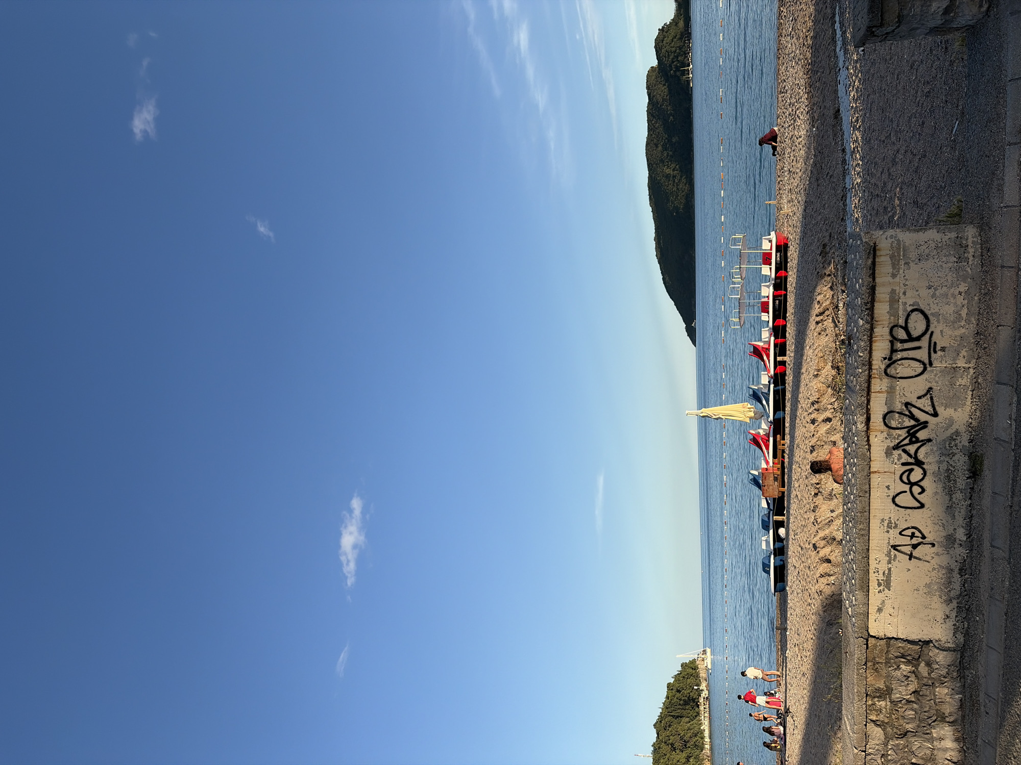 The image captures a sunny late afternoon scene at a beach, likely in Budva, Montenegro, given the location cue. The sky is a clear, deep blue with a few sparse, wispy white clouds.
On the right side, a calm, blue sea stretches towards the horizon, where it meets a lighter sky. A line of white buoys marks a swimming area in the water. A large, verdant mountain or peninsula, densely covered in trees, forms the coastline, extending into the sea on the far right.
A narrow strip of pebbly beach runs along the middle of the image, separating the sea from a concrete pathway or barrier on the left. Several people are visible on the beach. One individual, wearing a dark top, is seen bending down near the water's edge. Further down the beach, a small group of people is gathered near a line of paddle boats, some standing, some seemingly seated. The paddle boats are colorful, with prominent red, white, and blue sections. A yellow beach umbrella is also open on the sand near these boats. Another person is partially visible, lying on the sand closer to the concrete barrier.
The concrete barrier, running along the left foreground, features visible graffiti. The most prominent part of the graffiti appears to read "AD GORANA OIE" or similar, with the letters "OTB" clearly legible as a separate tag above it. The light suggests late afternoon or early evening, casting long shadows on the beach and indicating a warm, pleasant day. The overall atmosphere is one of leisure and relaxation at a popular coastal spot.