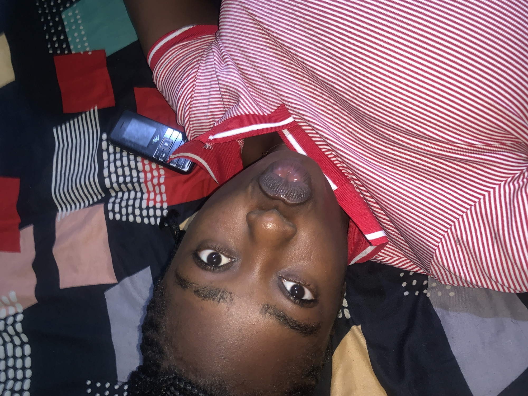 The image shows a close-up, upside-down view of a person lying on a bed with a patterned duvet. The person's face is centered, with their eyes open and looking upwards. They are wearing a red and white striped collared shirt. To the left of the person's face, a black mobile phone is visible, lying on the duvet. The duvet has a colorful geometric pattern with squares of red, black, light blue, and pink, interspersed with white dots. The background shows the edge of a pillow or blanket with a black and white stripe pattern. The overall scene appears to be a casual, indoor setting.