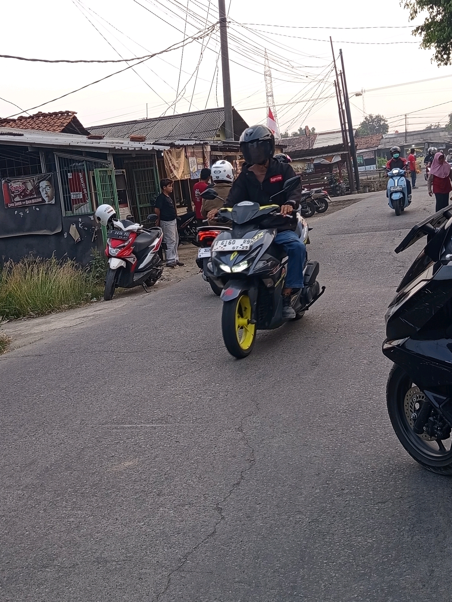 In Karawang, Indonesia, a motorcycle rider wearing a black helmet and jacket navigates a paved road. They are riding a black motorcycle with yellow accents, its headlights illuminated. Other motorcycles are visible on the road, including a red and white one parked on the left and a light blue one further ahead. Several people are seen near buildings lining the street, with some wearing helmets and others not. The environment suggests a commercial or residential area with overhead electrical wires crisscrossing the sky, and the time appears to be late afternoon or early evening given the soft, diffused light.