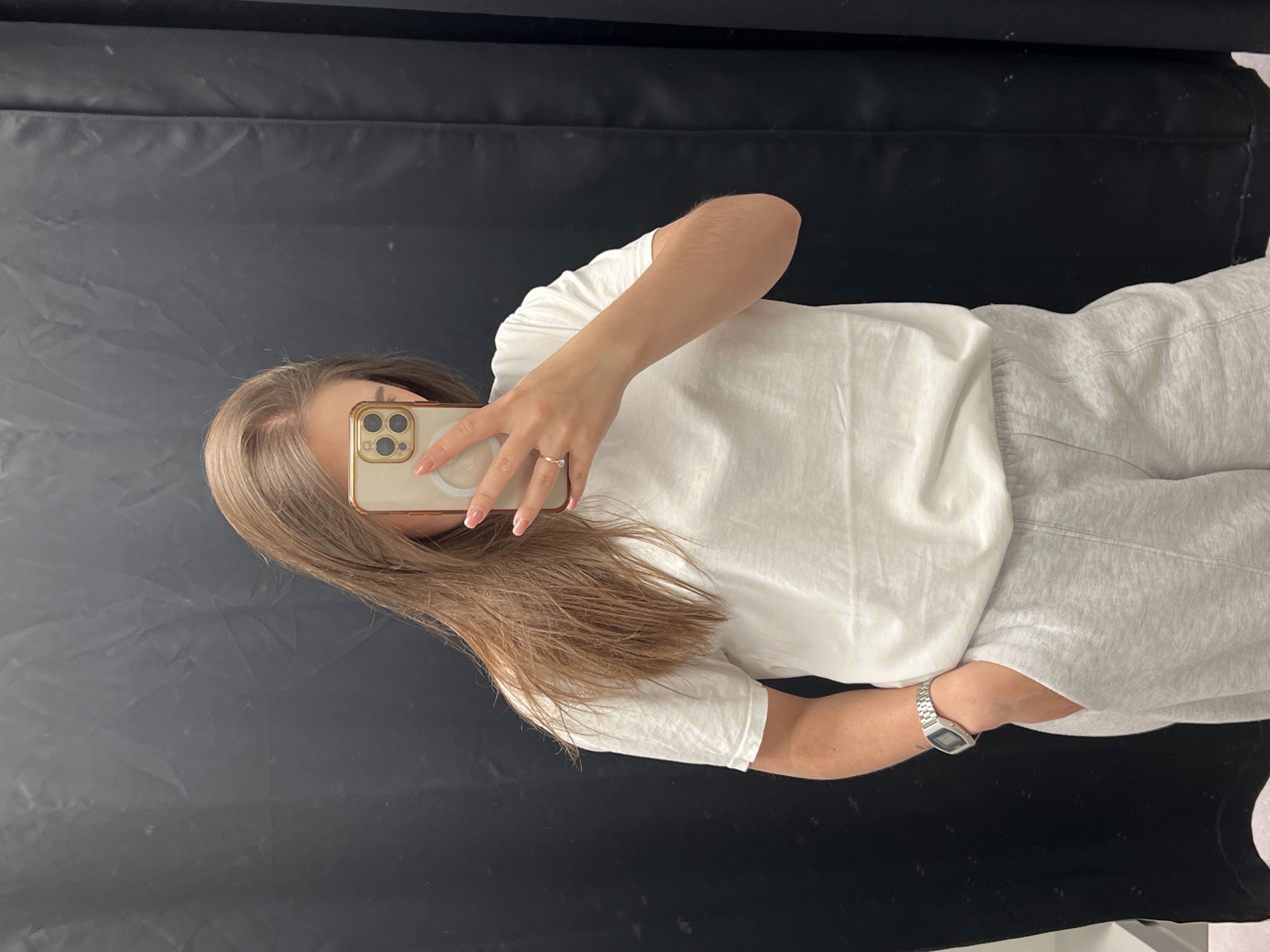 A person takes a selfie in front of a black backdrop. The person has medium-length brown hair and is wearing a white t-shirt, light grey sweatpants, and a silver watch on their left wrist. They are holding a smartphone with a gold-colored case, obscuring their face. A ring is visible on the ring finger of their left hand. The location of the image is unknown.