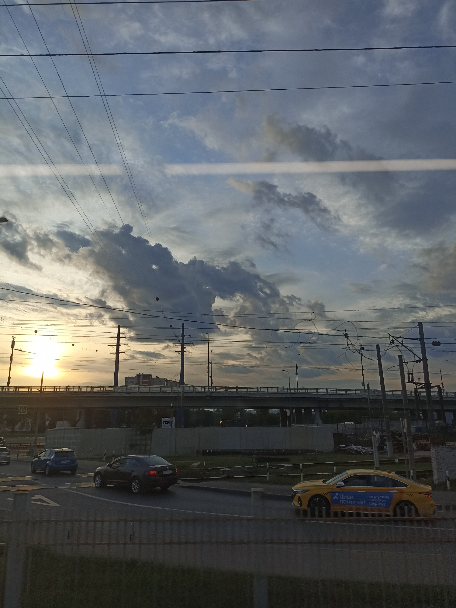 A wide shot captures a city intersection during sunset, with a dramatic sky filled with clouds and wires overhead. In the foreground, a yellow taxi with Russian advertisements drives on the road. A dark sedan and a blue SUV are also visible, along with a pedestrian crossing and a traffic light. The background features an elevated highway and urban buildings, while power lines and utility poles crisscross the scene. The sun, low on the horizon, casts a warm glow, illuminating the clouds and the cityscape.