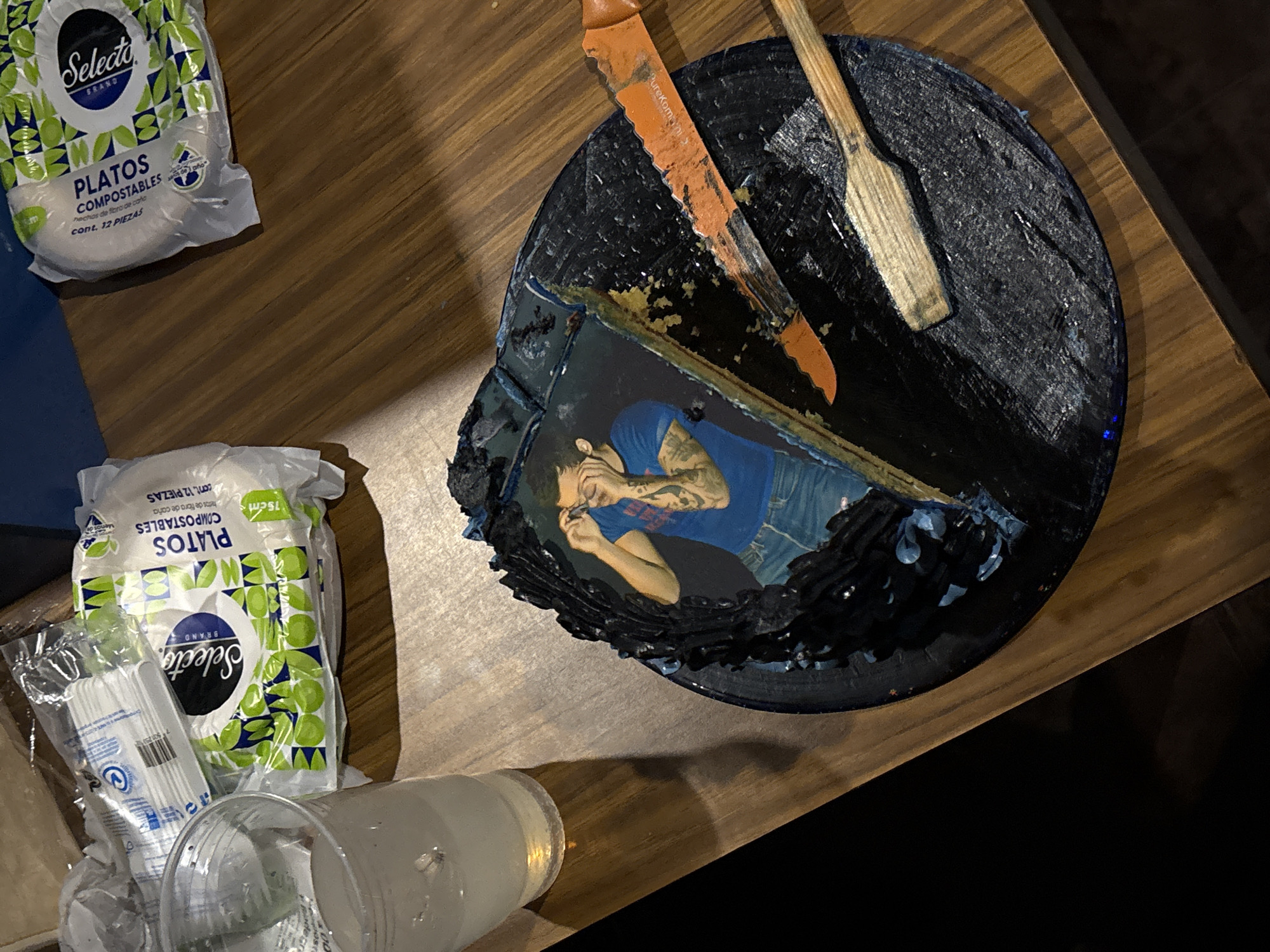 The image shows a partially eaten cake on a wooden table, accompanied by disposable tableware and plastic cups. The cake is round with dark blue frosting, and a slice has been cut. The cut slice reveals an image of a tattooed man on the inside. A knife and a wooden utensil rest on top of the cake. Next to the cake are a package of Selecto Brand compostable plates, a package of Selecto Brand plastic cutlery, and several plastic cups. The location appears to be indoors. There is no other information in the image to support a location in San Bernardino Tlaxcalancingo, Mexico.