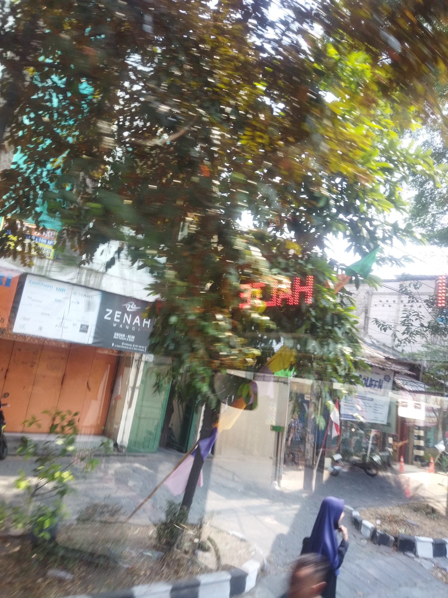 In Surakarta, Indonesia, an outdoor scene unfolds with a notable red LED sign displaying "3.3AH" amongst lush green foliage. To the left, a storefront with orange doors is visible, featuring a sign with text including "ZENAH WANGI" and "GROSIR & ECER". Another sign above it is partially obscured by leaves but shows text like "iberchem uzi". A person wearing a blue hijab stands near the curb, facing away from the camera, with another person's head in the foreground. The ground appears to be a paved area with some rubble and vegetation. A traffic cone is visible in the background. The image is blurred, suggesting it was taken from a moving vehicle.