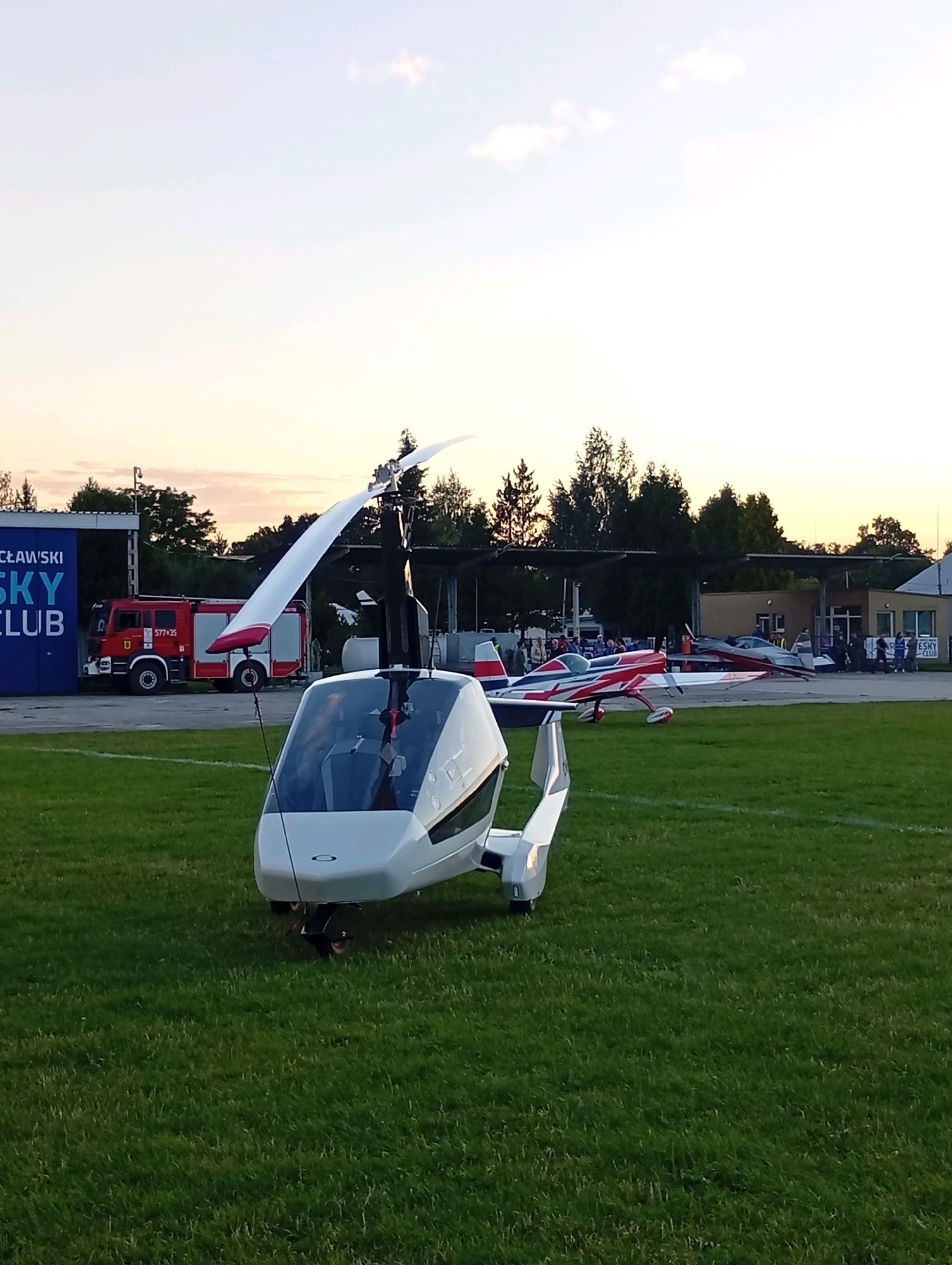 The image depicts an outdoor scene at an airfield during what appears to be late afternoon or early evening, evidenced by the soft, warm light and pastel colors in the sky. In the foreground, a white autogyro with a prominent rotor is parked on a grassy field. The autogyro has a sleek, modern design with a transparent canopy. Behind it, several other aircraft are visible, including a red and white acrobatic plane.
Further in the background, there is a building complex with covered areas, possibly hangars or a club house. A red fire truck with "577+35" displayed on its side is parked near a blue structure that has text which partially reads "ŁAWSKI SKY CLUB". Several people are gathered in small groups around the airfield, some near the buildings and others near the aircraft. The environment is characterized by manicured grass, trees in the distance, and a generally clear sky with a few wispy clouds. The overall impression is one of an organized aviation event or a general aviation airport.