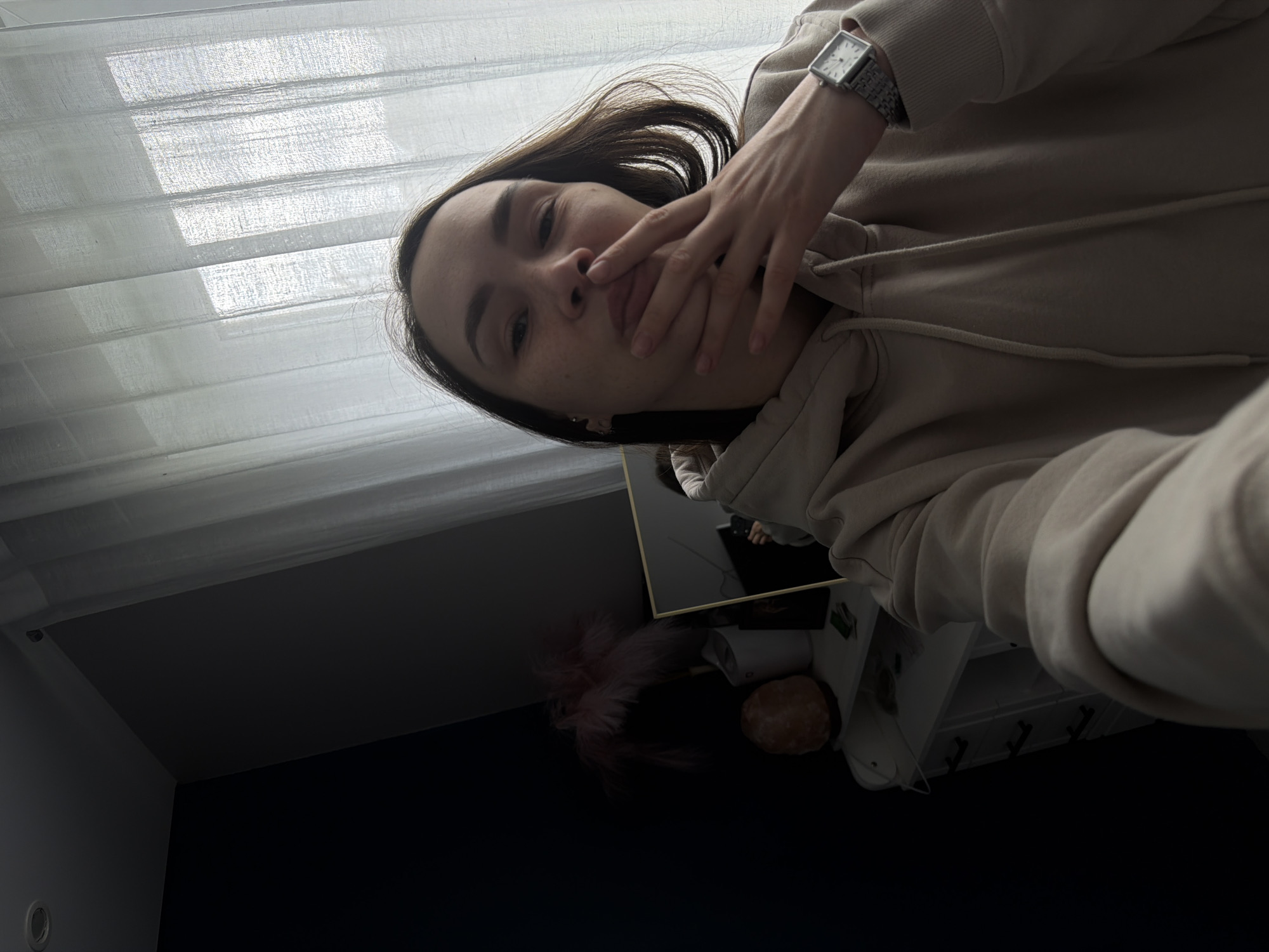 A person is taking a selfie indoors with a silver-colored square-faced watch on their left wrist, wearing a beige hoodie, and has their left hand covering their mouth. The background includes a window with sheer white curtains, a dark-colored wall, and a blurred reflection in a mirror showing another person. A small table with shelves is also partially visible. The location is reported to be Chaikovsky, Russia.