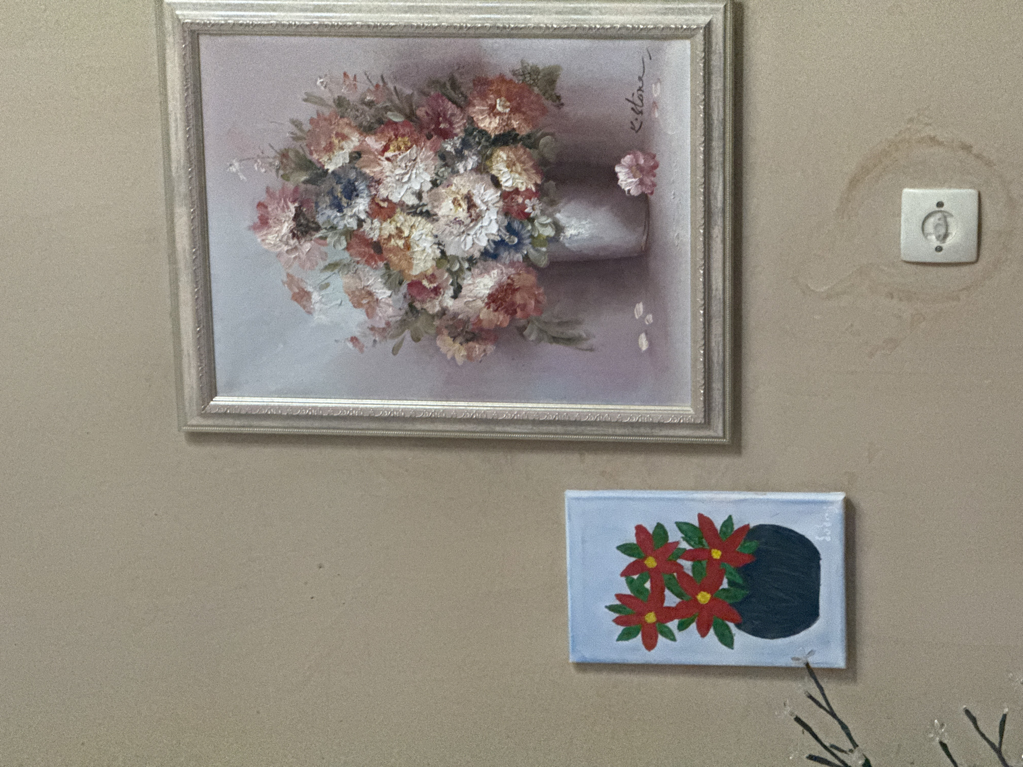 The image shows a section of a wall in an indoor setting. The wall is painted in a neutral beige tone and has a circular water stain around a white electrical outlet. Two framed paintings hang on the wall. The upper painting is larger and depicts a bouquet of flowers in a vase, rendered in soft pastel colors within a silver frame. The lower painting is smaller and shows a still life of red flowers with green leaves against a light blue background with a dark round object. The bottom edge of a decorative floral arrangement with thin black branches and clear embellishments is visible. It is not possible to determine the location in Tbilisi, Georgia based on the content of the image.