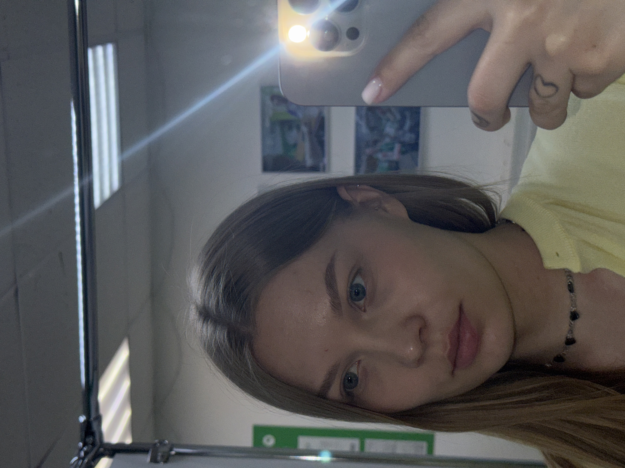 A young woman is taking a selfie in a mirror. The mirror shows a room with two framed pictures hanging on the wall and a fluorescent light fixture. The woman is wearing a yellow top and a necklace with dark beads. She has a small heart tattoo on her finger. The location cannot be determined, as no information is available that supports the exact location.