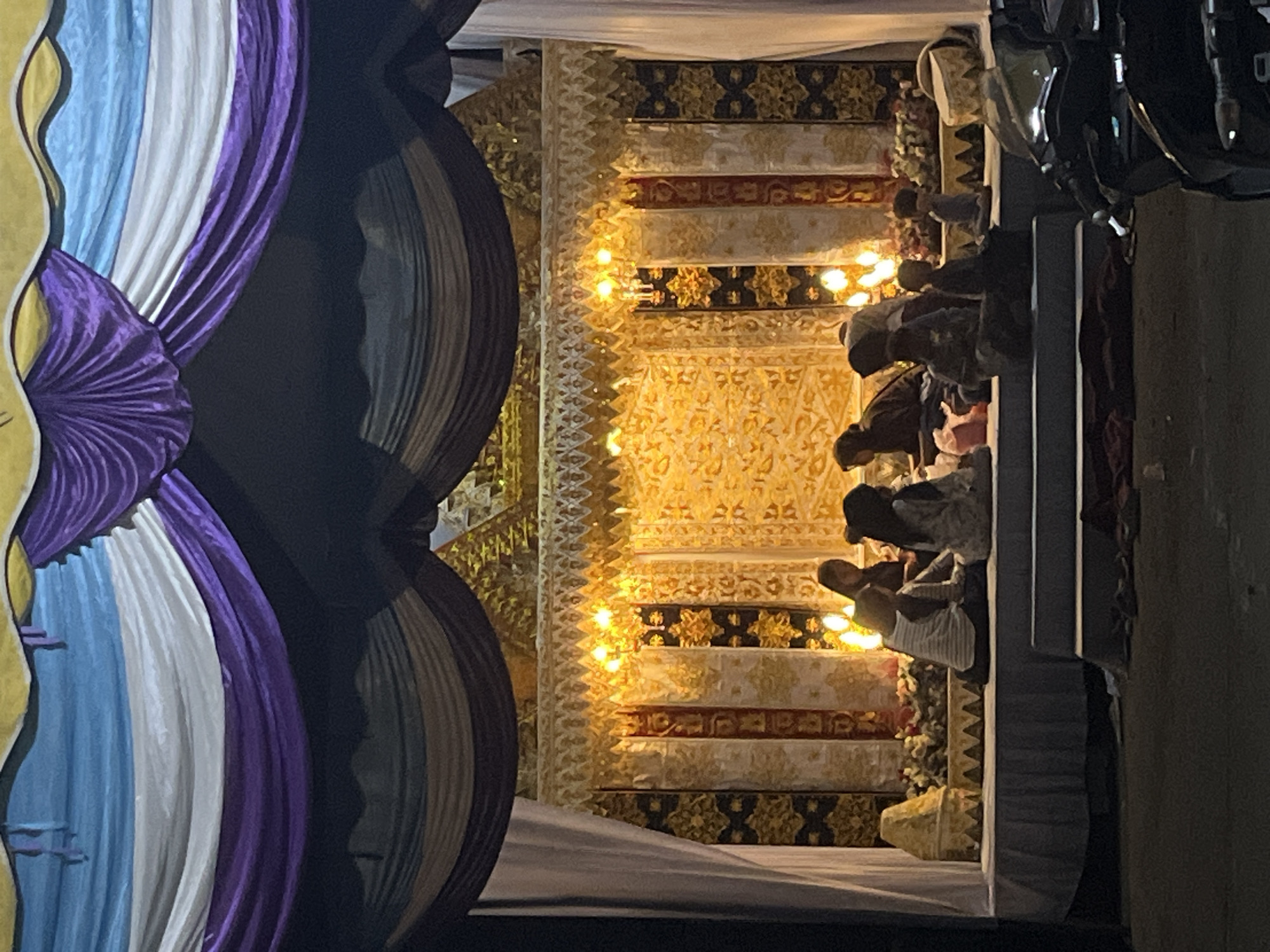 This image captures a vibrant and ornately decorated scene, likely an event stage or venue, illuminated by artificial lighting at night.
On the left side, large fabric drapes in striking shades of purple, white, and light blue are prominently displayed, forming sweeping folds as part of the decoration. A section of golden material with an organic, wavy design is also visible on the far left edge.
The central and background area features an elaborate golden backdrop, rich with intricate geometric and floral patterns. This backdrop is composed of alternating bands of solid gold designs, sections of dark fabric with golden motifs, and cream-colored panels with subtle patterns. Numerous small, bright, warm-toned lights are strung across the golden backdrop, casting a festive glow and highlighting the intricate details.
On the right side of the frame, several individuals are gathered, appearing to be seated or kneeling on a low platform or the ground. They are seen from behind or the side, dressed in various modest attire, with many wearing head coverings. Their postures suggest they are observing an event or participating in a ceremony on the decorated stage. Pillars or structural elements are also visible in the foreground on this side.
The setting, decorative style, and the gathering of people, combined with the location context of Lhokseumawe, Indonesia, suggest this is a cultural event, celebration, or religious gathering. The scene is entirely lit by artificial light, indicating it is nighttime. There is no discernible text in the image.