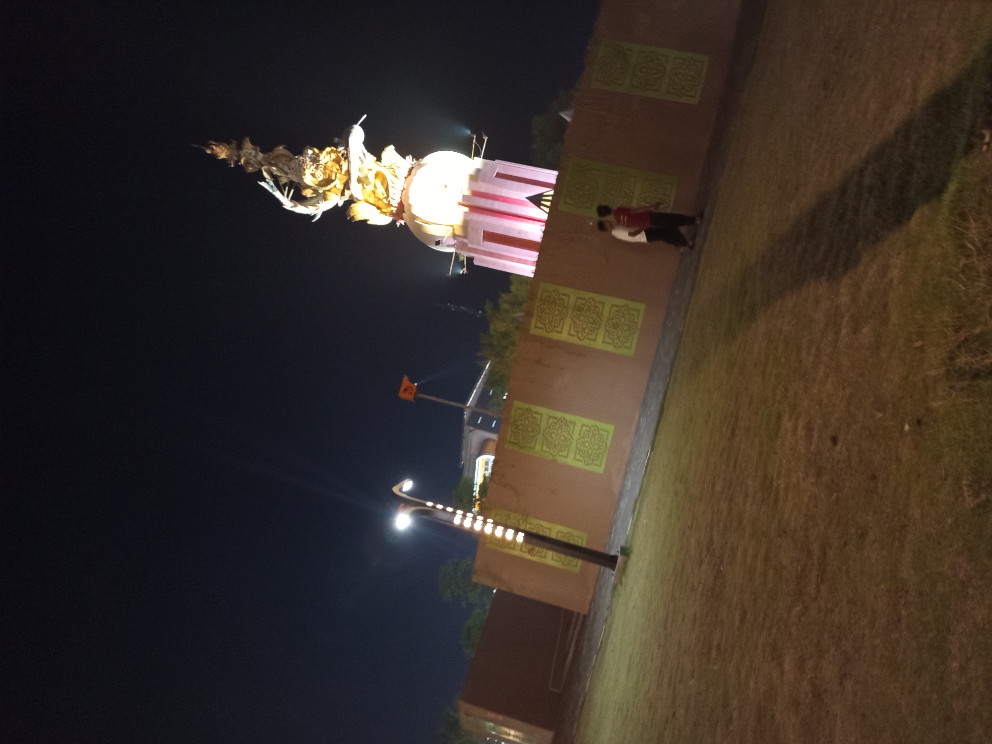 In Sidoarjo, Indonesia, at night, a large, illuminated monument with golden, intricate sculptures atop a circular base and a pink-accented tower stands against a dark sky. A wall, adorned with green, repeating decorative patterns, encircles the monument. In front of the wall, a well-maintained grassy area slopes down to the foreground. A contemporary lamppost with multiple lights is situated to the left of the monument. Two individuals are visible walking along the pathway behind the wall.