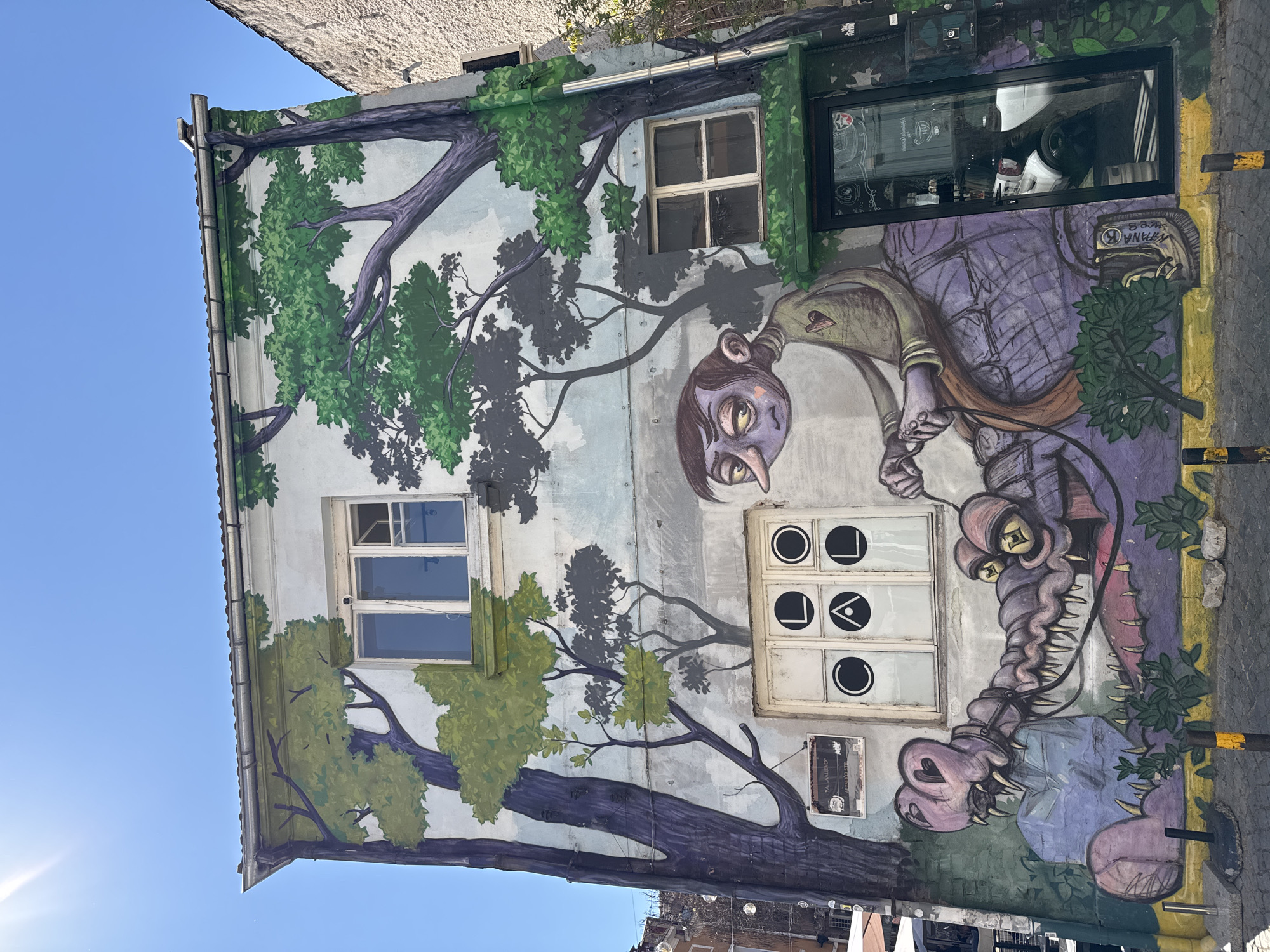 The image shows the facade of a building in Plovdiv, Bulgaria, covered in a large mural. The mural features trees, a grotesque cartoon-like character, and a monster-like figure. There are several windows on the building, some of which are integrated into the mural design. The sky is clear and blue in the upper left corner of the image. The scene appears to be outdoors, and the building is located in an urban environment.