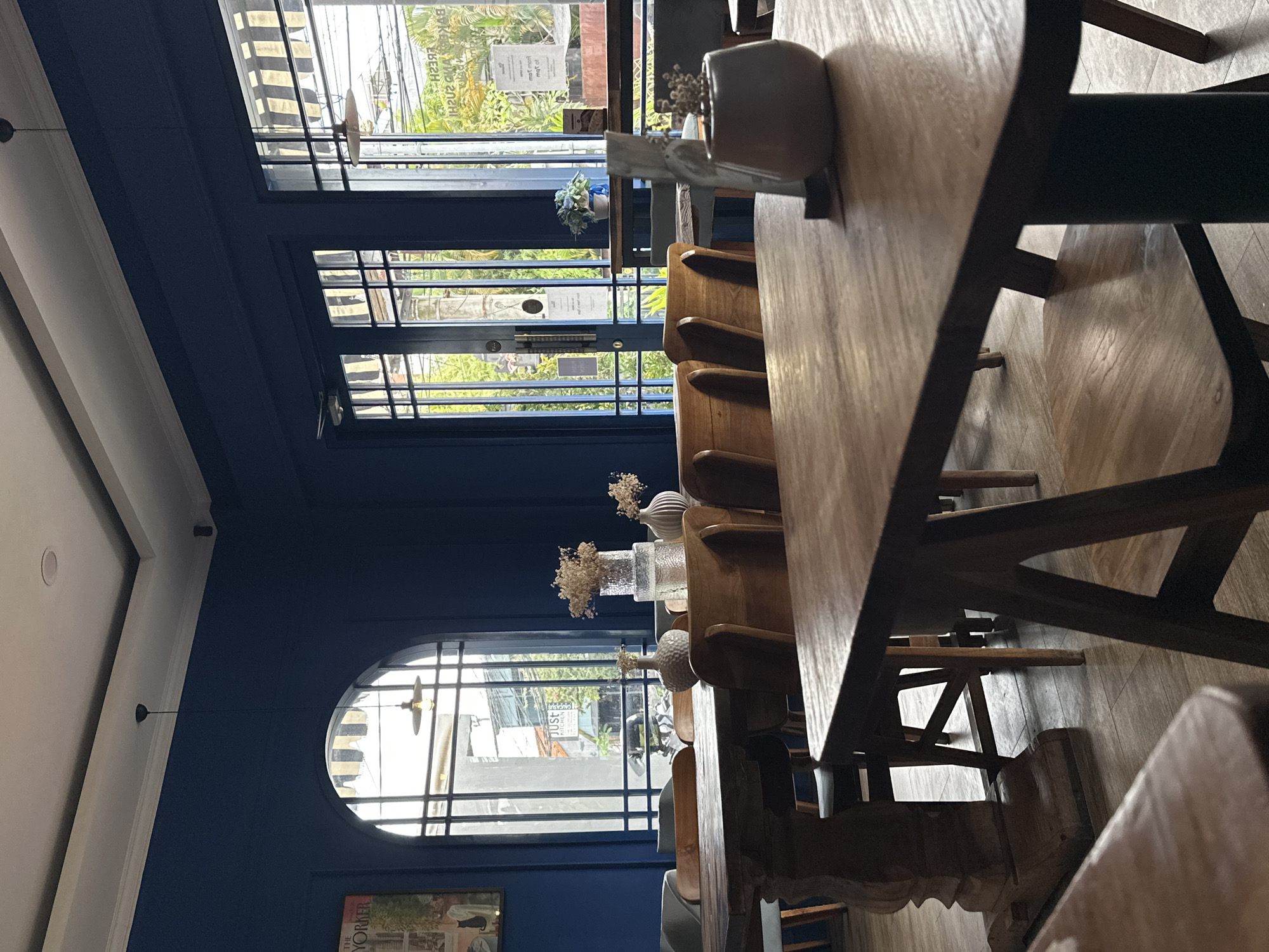 The image captures the interior of a restaurant or cafe, potentially in North Kuta, Indonesia, judging by the tropical foliage visible through the windows. The dominant feature is a long, wooden table with several chairs arranged along its side. The table is decorated with small vases containing dried flowers. The room is painted a deep blue color. Large windows, including one that is round at the bottom, provide natural light. A picture frame with a print is visible near the bottom of the round window. The image composition is somewhat unusual, with the table and chairs presented in a diagonal perspective. No people are visible.