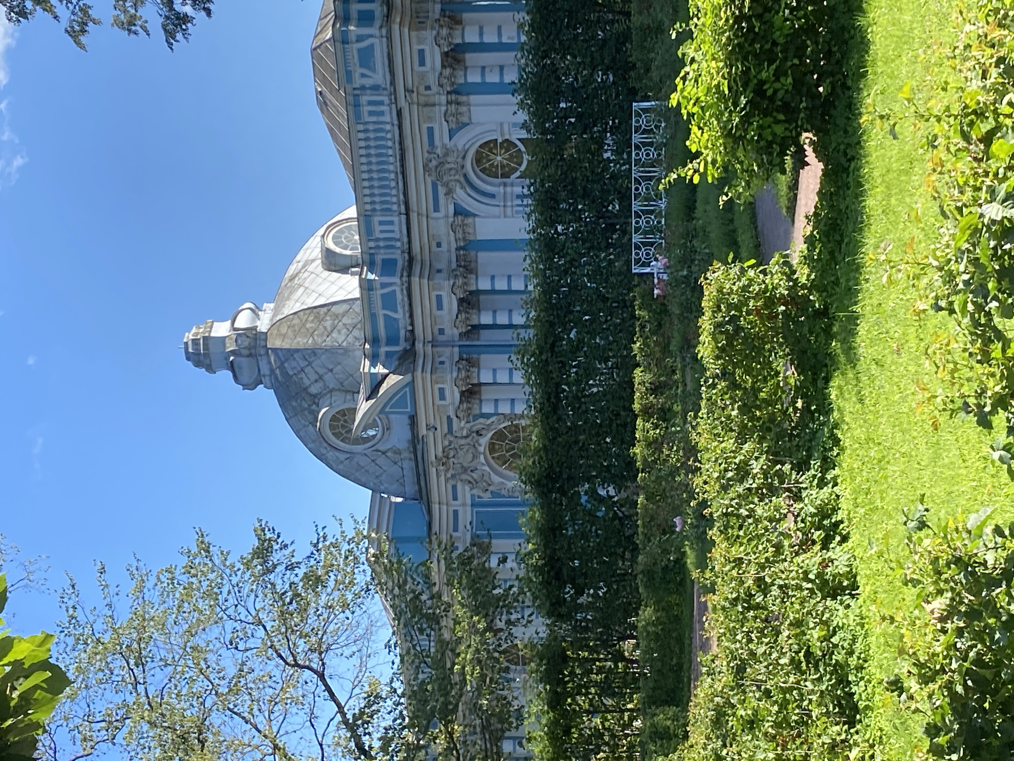 The image captures a sunny day in the city of Pushkin, Russia, showcasing a portion of a grand architectural structure behind a manicured garden. The building, with its distinctive blue and white facade, features a prominent dome and ornate architectural details including columns and decorative windows. The garden is lush and green, with neatly trimmed hedges and trees. In the foreground, a vibrant green lawn is visible, interspersed with shrubs. A white decorative fence and a pathway are also present within the garden. The sky is a clear, bright blue, indicating fair weather. The scene is tranquil and well-maintained, suggesting a public park or historical site.