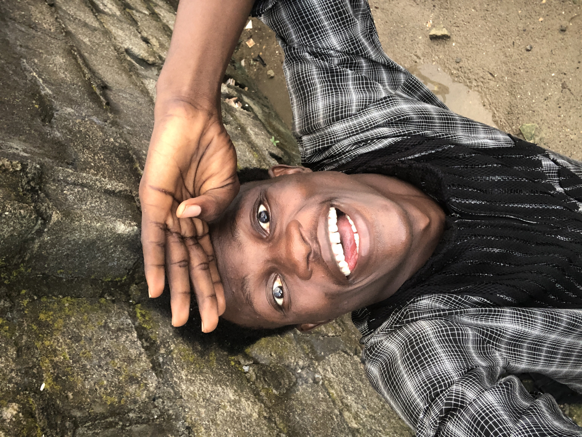 A young Black male, with dark skin and short, dark, curly hair, is captured in a selfie taken from a high angle. He is smiling broadly, with his mouth open and tongue visible, and his eyes are wide open and looking directly at the camera. His right hand is raised to his forehead, obscuring a portion of his hairline. He is wearing a black and white plaid shirt over a black garment with a textured pattern. The background consists of a rough, grey stone wall, appearing weathered and partially covered in moss, to the left of the frame. To the right, the ground is a mix of damp sand and some debris, possibly indicating an outdoor, possibly unpaved area in Ado, Nigeria.