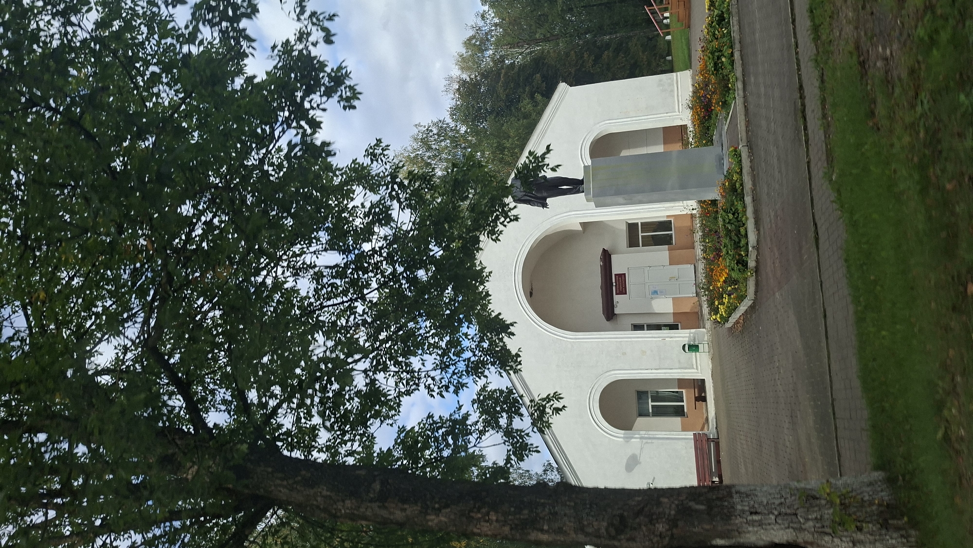 The image depicts a public building with a white facade, featuring large arched entrances. In front of the building, a statue of a man stands on a stone pedestal, surrounded by a flower bed. To the left of the statue, there is a red park bench and a green trash can. The ground in front of the building is paved with bricks. Trees and grass are visible in the foreground and background. The sky is partly cloudy. The building appears to be located in a town or village setting, possibly within the Бягомльскі сельскі Савет in Belarus.