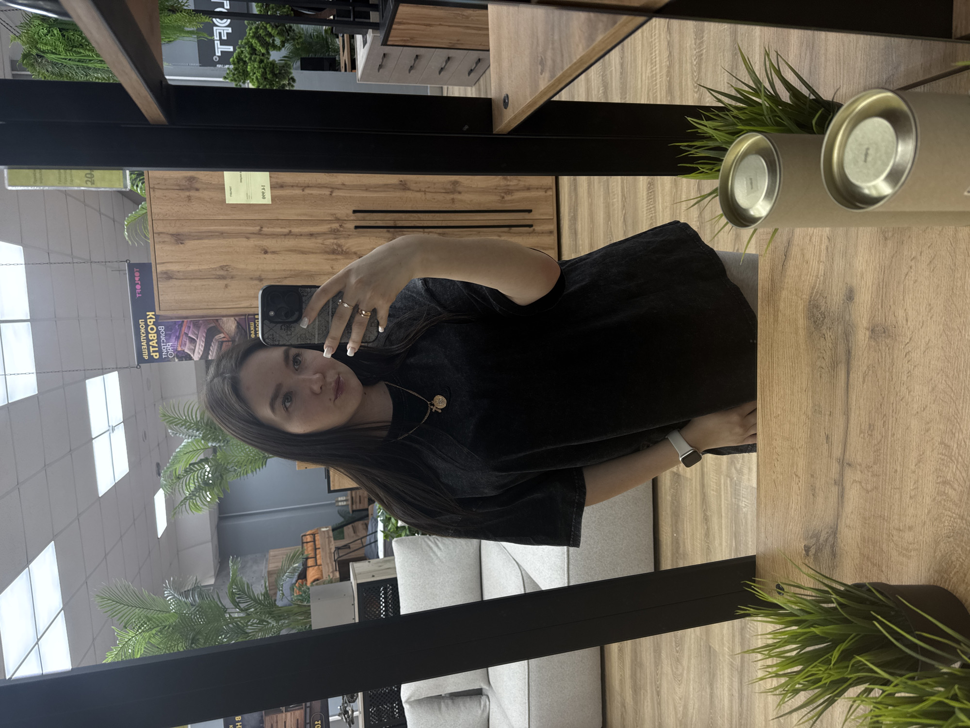 Here's a detailed description of the image:
**Scene:** The image appears to be a selfie taken in a furniture store, possibly in Chaikovsky, Russia, based on the Cyrillic text in the reflection. The photo is being taken in front of a mirror that is part of a furniture display.
**People:** A young woman is holding a smartphone and taking the selfie. She has long brown hair, a gold necklace, and is wearing a black t-shirt. Her hand is visible, wearing a silver smartwatch.
**Subjects/Objects:**
* **Furniture:** The display includes cabinets with wood grain finishes and a light grey sofa or sectional.
* **Plants:** There are potted plants (likely artificial) on top of the furniture.
* **Signage:** A sign with Cyrillic text is visible in the background.
* **Other:** There are also additional furniture pieces and decorative elements visible in the background through the mirror's reflection.
**Actions/Interactions:**
* The woman is taking a selfie using her smartphone in front of a mirrored furniture display.
**Environment:**
* **Indoor:** The setting is an indoor store environment, likely a furniture showroom.
* **Lighting:** The area appears to be well-lit with overhead lighting.