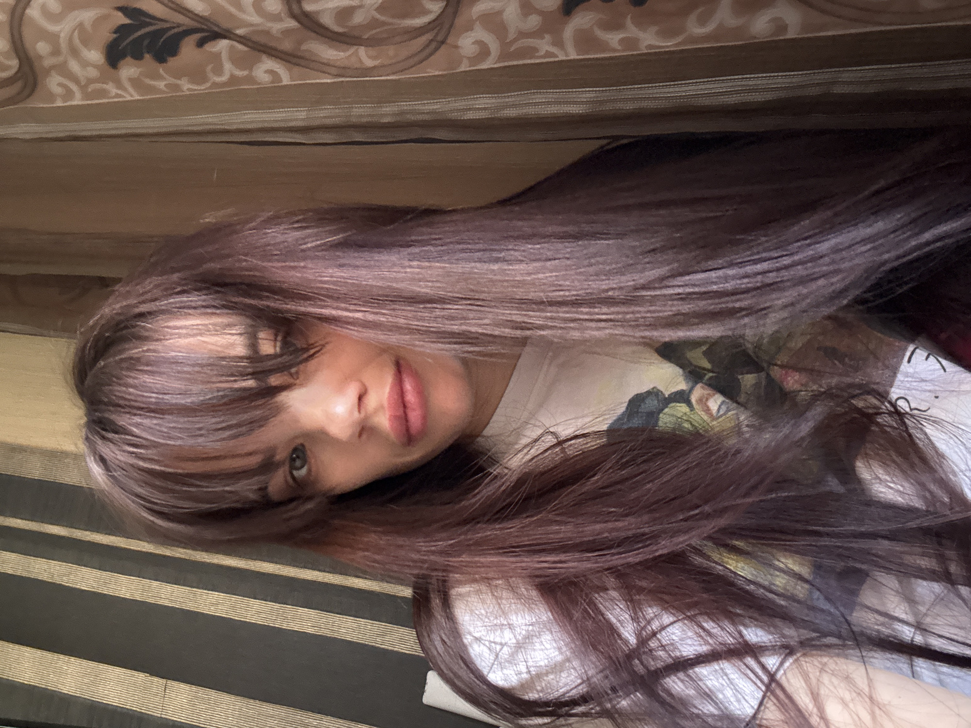 A young woman with long, dark hair and bangs is lying down, looking up at the camera. The background consists of striped fabric and floral fabric. The location is not discernible from the image.