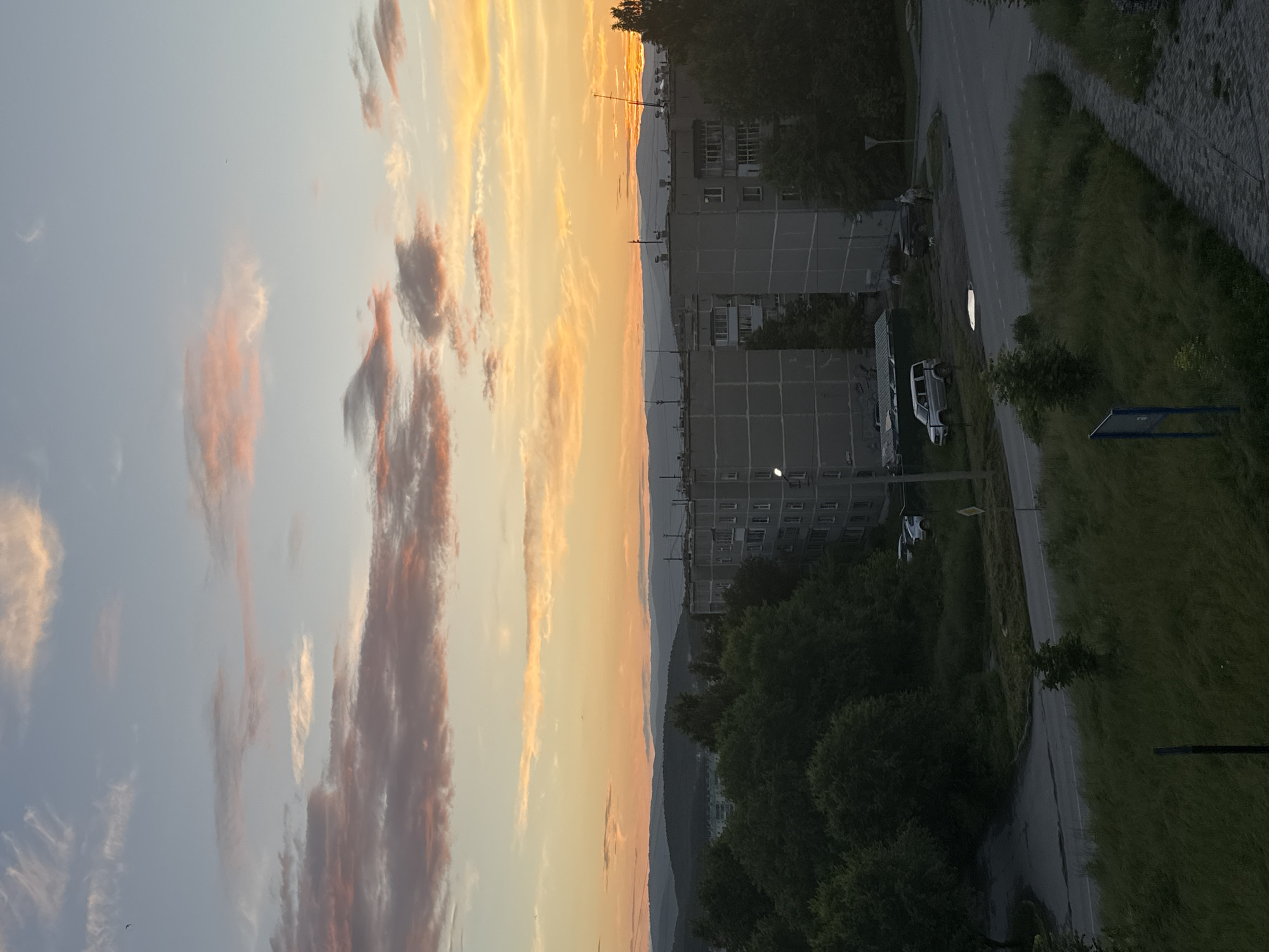 The image depicts a sunset over a residential area in Miass, Russia. The sky is filled with hues of orange, pink, and yellow, with scattered clouds. In the foreground, there are apartment buildings and trees. The buildings are concrete structures with multiple floors and balconies, characteristic of Soviet-era architecture. A road with cars is visible in front of the buildings, with a paved walkway bordering the grassy area. The landscape beyond the buildings features rolling hills and mountains, also shrouded in the soft light of the setting sun. The overall scene conveys a sense of calm and quiet evening in a Russian city.
