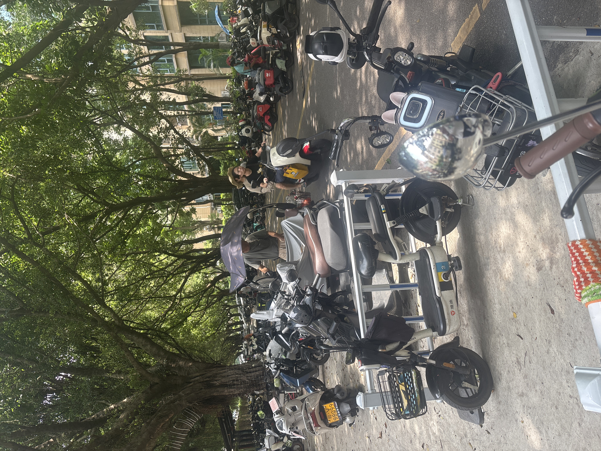 In Nanhai District, China, an outdoor scene depicts a densely packed parking area for electric scooters and motorcycles. The ground is a mix of concrete and asphalt. Tall, mature trees with lush green foliage provide dappled shade over the entire area, with several modern, multi-story apartment buildings visible in the background. A person in a green shirt stands under a dark umbrella, seemingly tending to a scooter covered with a grey tarp. Further into the background, a young couple rides a white electric scooter, the passenger holding a yellow bag. In the foreground, several electric scooters are parked. One white scooter in the mid-ground has a basket with items inside, a license plate with "98099" visible, and Chinese characters on its body. Another scooter in the foreground is mostly obscured by a railing and features a silver mirror reflecting the surroundings. The overall impression is a busy urban environment with a focus on personal electric transportation.