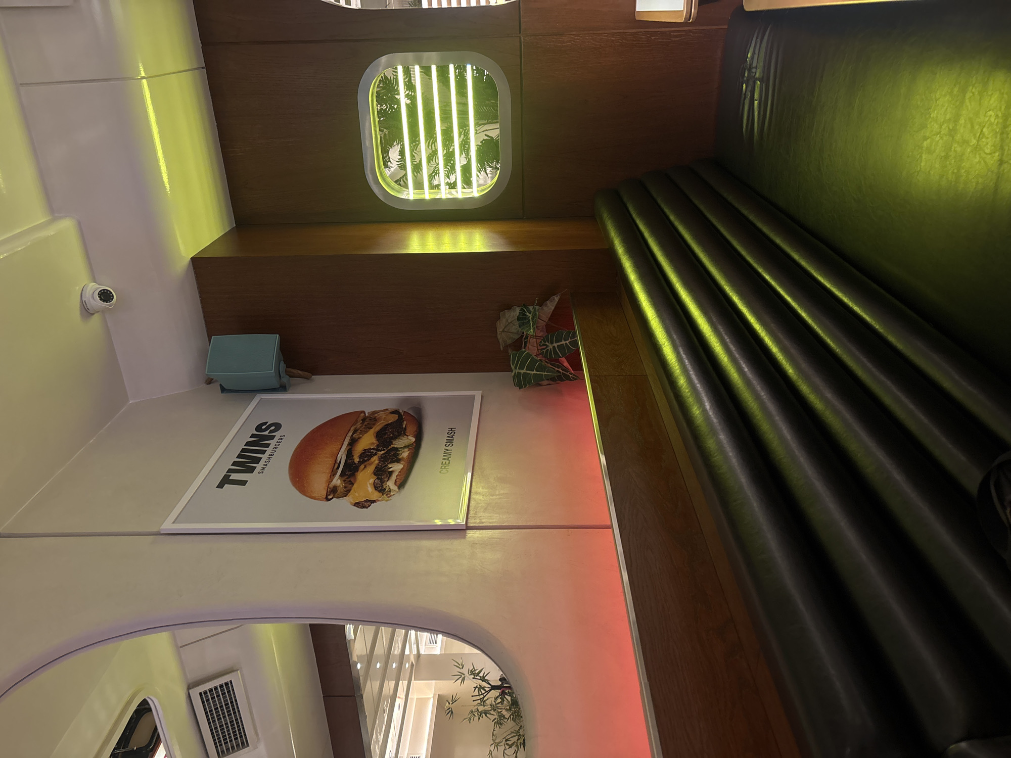 This image captures the interior of a modern burger restaurant or diner, located in Yerevan, Armenia. The scene is devoid of people, focusing on the establishment's decor and ambiance.
The primary elements include a long, dark green or black leather booth with horizontal tufting, illuminated by a yellowish-green light source, creating a warm and somewhat moody atmosphere. Adjacent to the booth, a section of dark wooden paneling extends upwards, featuring a distinctive porthole-shaped window with vertical green neon lights that highlight lush green foliage visible outside.
On the opposite wall, a white surface is prominent, upon which a white-framed poster is displayed. The poster showcases a close-up image of a large, appetizing burger, with the text "TWINS SMASHBURGERS" at the top and "CREAMY SMASH" printed along the bottom edge of the burger. Below the poster, a light blue, rectangular object rests on a shelf. Higher up on the white wall, a small white security camera is mounted.
Below the porthole window, on a lower shelf, a decorative item resembling a patterned fabric or wrapper, featuring green and red tropical leaves, is placed. The lower portion of the image reveals a reflective surface, possibly a mirror or polished floor, showing inverted reflections of the interior, including a white air vent and some decorative bamboo-like plants.
The lighting is predominantly artificial, with the yellowish-green hues dominating the space and a distinct green from the neon window. This, combined with the stylish interior design elements like the porthole and dark wood, suggests an evening setting or a design choice emphasizing a contemporary, possibly themed (e.g., retro-futuristic or nautical) dining experience.