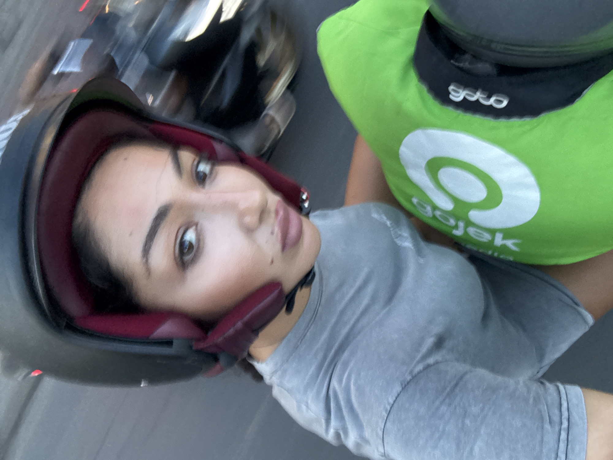 A woman wearing a dark grey helmet with maroon padding takes a selfie while on a moving motorcycle. Beside her is a person wearing a bright green vest with the Gojek logo, indicating a ride-hailing service common in Southeast Asia. The image appears to be taken in motion, likely in an urban environment. Given the Gojek logo, and the prompt specifying Kuta Selatan, Indonesia, the location is presumed to be in Kuta Selatan, Indonesia.