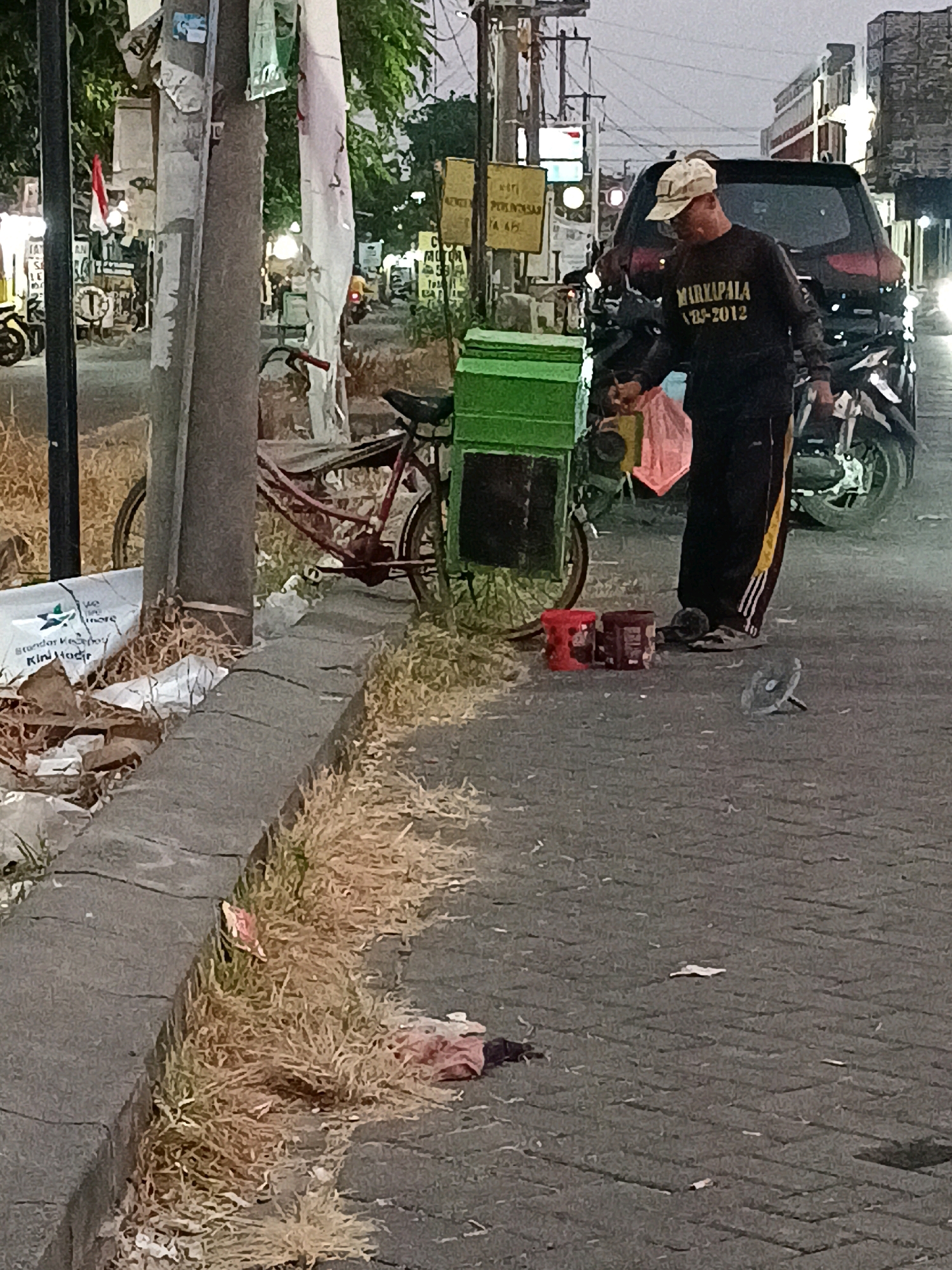In Kletek, Indonesia, a man wearing a light-colored cap and a black long-sleeved shirt with the text "MARKAPALA 1995-2012" printed in yellow stands near a green cart. He is holding a yellow object in his right hand. The cart is positioned next to a red bicycle with a black seat and a silver kickstand. The scene takes place on a paved street, with dry grass and debris along the roadside. In the background, a dark-colored SUV is parked, and a motorcycle is visible to its right. Other elements in the environment include streetlights, signs, and buildings lining the street. There are two red cylindrical containers and a gray circular object on the ground near the man.