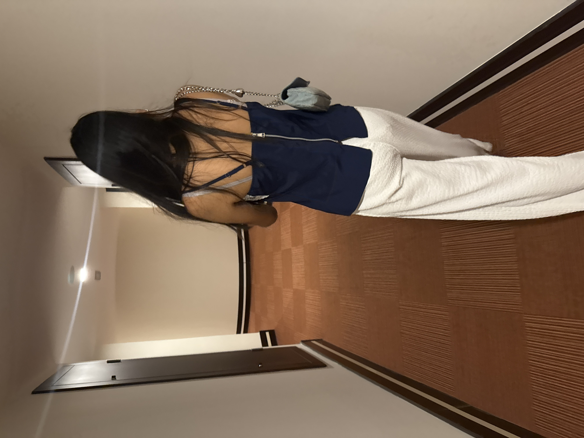 The image shows a person walking down a hotel hallway. The hallway has a patterned brown carpet and white walls with a dark trim. There are doors on the left side of the hallway. The person is wearing a dark blue sleeveless top with a zipper down the back and white wide-leg pants. They are carrying a light blue purse with a chain strap. The image appears to be taken from behind the person, looking down the hallway. The location is not specified as being in Aksu, Turkey.