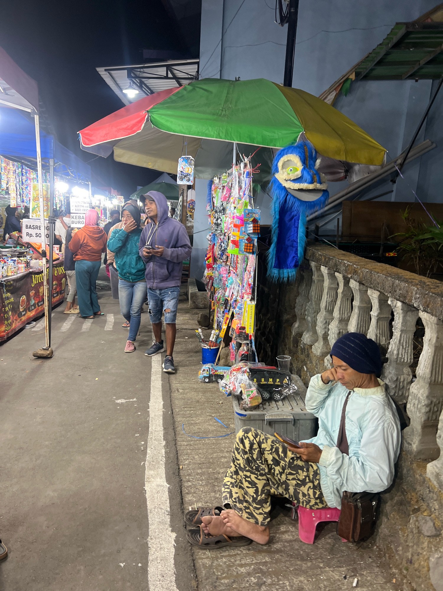 An outdoor street market stall, potentially located in Windusari, Indonesia, is depicted at night. A vibrant, multi-colored umbrella, red, green, and yellow, shades a variety of merchandise displayed on racks, including toys and trinkets. A large, blue furry monster costume with a yellow and white face is also present. To the right, a person wearing a dark blue beanie, a light blue jacket, and camouflage pants is seated on a pink stool, engrossed in their mobile phone. They are resting their elbow on a grey storage container. Adjacent to the stall, several individuals are walking along the paved pathway, some carrying bags. The ground is paved, with a white line marking a boundary. The background features a dark wall and structures suggestive of an urban setting.