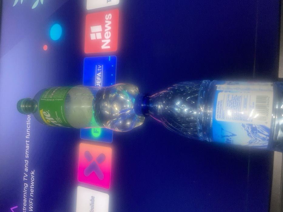 Two plastic bottles are joined at their mouths, positioned horizontally in the center of the image. One bottle contains a greenish liquid, likely Sprite, while the other holds clear water. These bottles are lying in front of a television screen displaying various app icons like News, YouTube, and JEFA.tv, with a blue and purple gradient background. Text on the screen mentions streaming TV, smart functions, and WiFi networks.